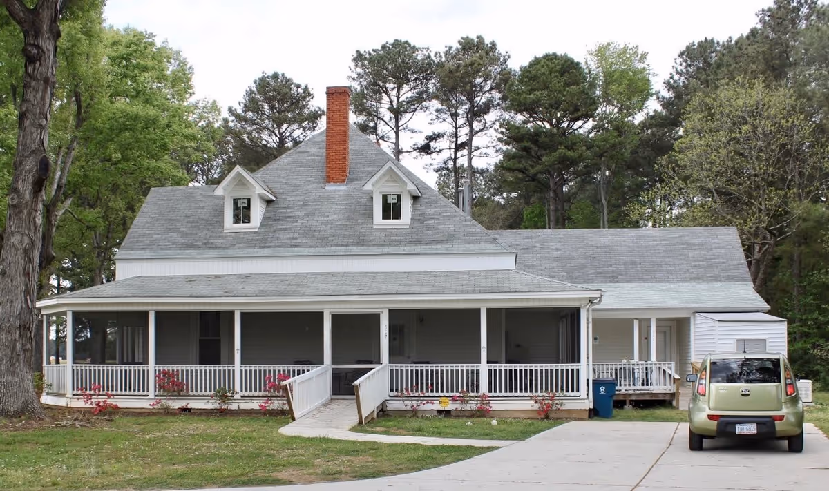 Front exterior view of a two-story house with a large covered porch, white railing, and a gray roof with two dormer windows and a red brick chimney. There is a green car parked on the driveway to the right, surrounded by trees and greenery.
