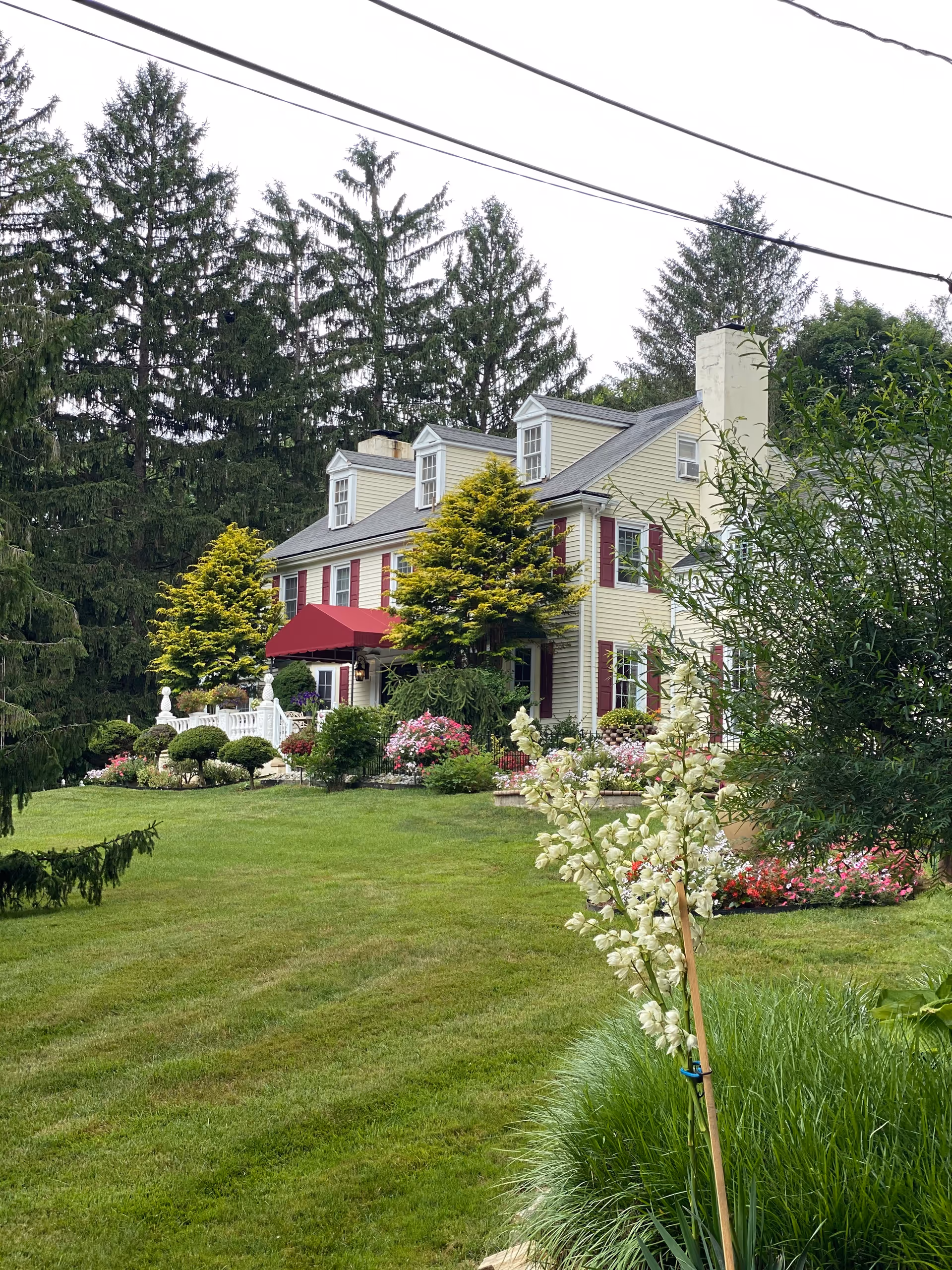 A large, two-story colonial-style house with cream-colored siding and red shutters, surrounded by a well-maintained lawn and various flowering plants and shrubs. There are tall evergreen trees in the background and a red awning over the front entrance.