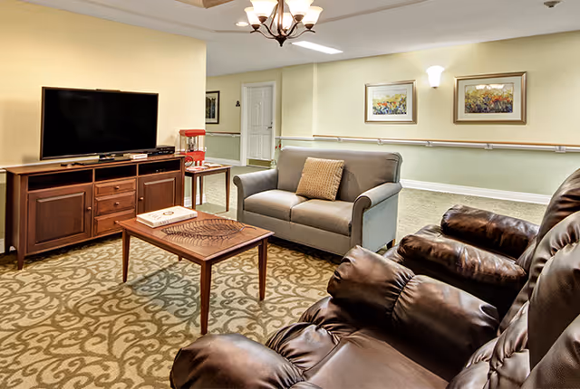 A comfortable senior living common room with leather recliners, a loveseat, coffee table, and a TV on a wooden console.
