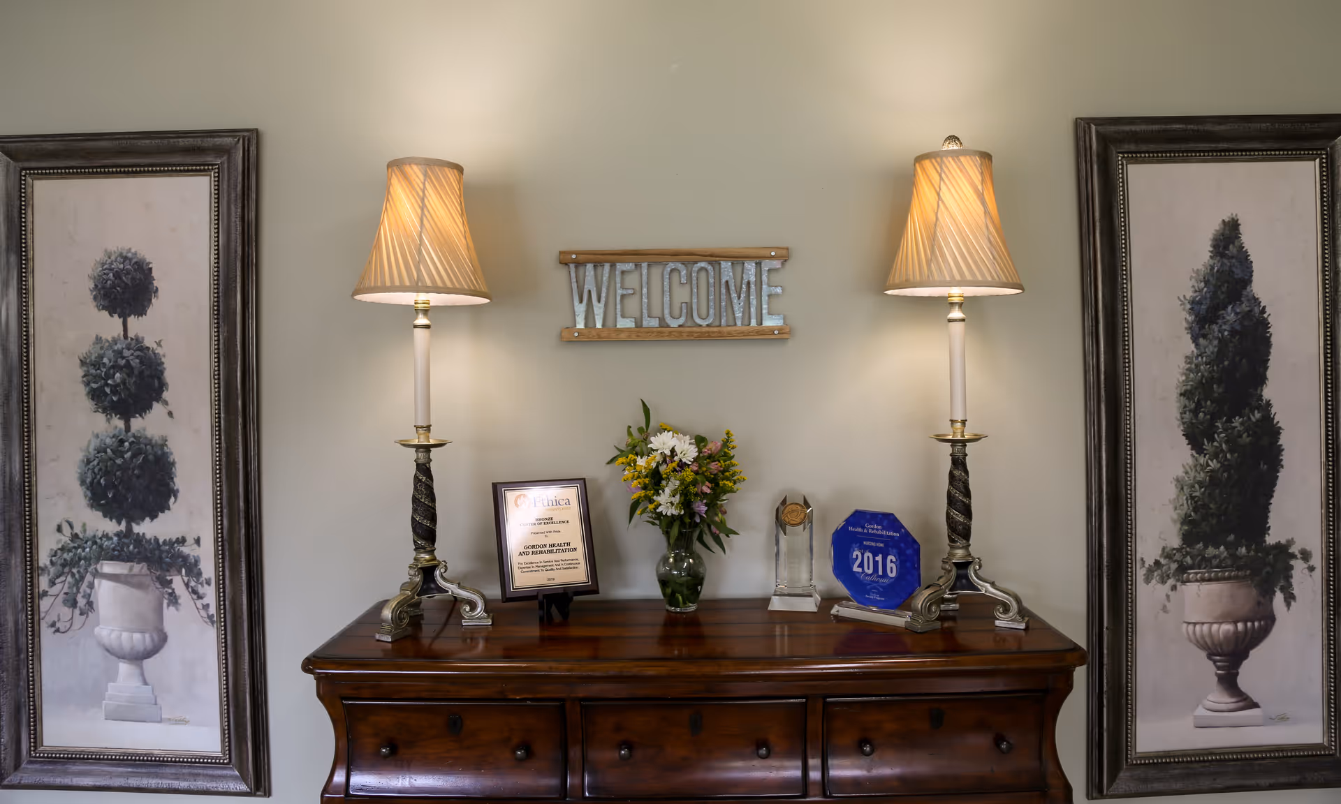 Wooden console table with two lamps, a welcome sign, flowers, and framed art in a lobby.