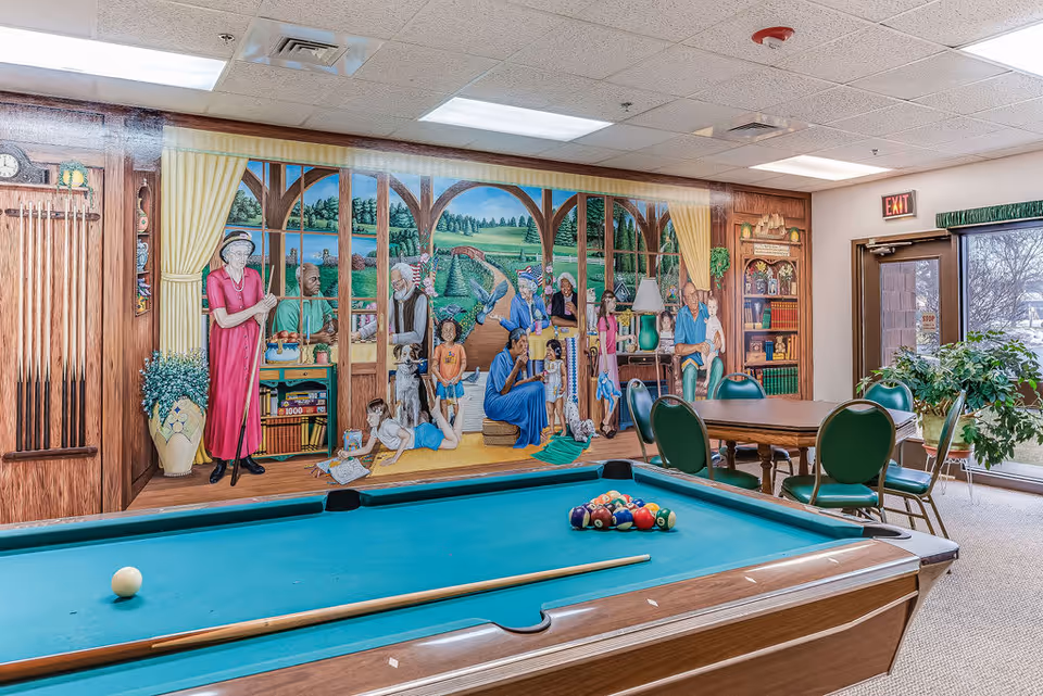 A recreational room with a pool table in the foreground and a mural on the wall depicting diverse elderly people and children in a garden setting. There is a table with green chairs near a glass door leading outside, and some plants are placed near the door.