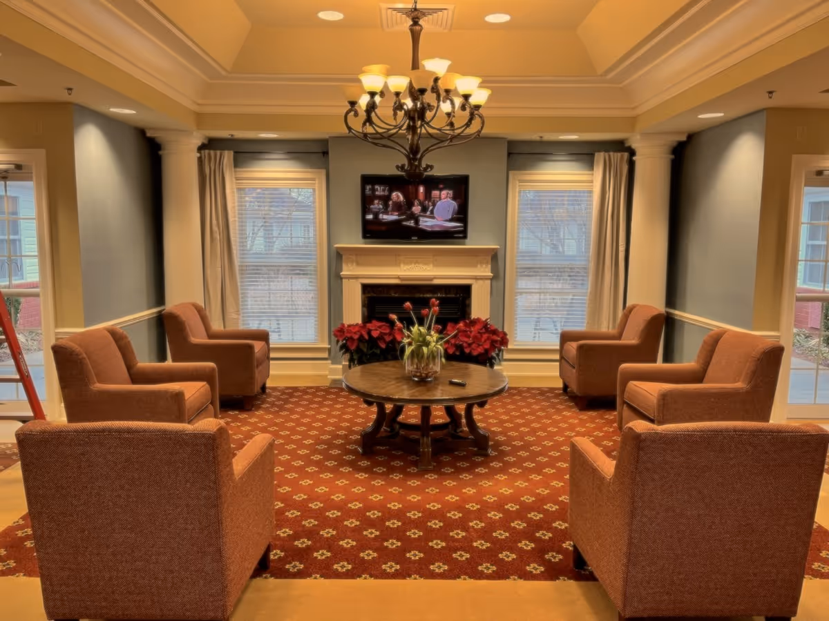 A cozy living room area with six brown upholstered armchairs arranged in a circle around a round wooden coffee table with a glass vase of flowers. The room features a red patterned carpet, a chandelier hanging from the ceiling, a fireplace with a TV mounted above it, and two large windows with curtains on either side of the fireplace.
