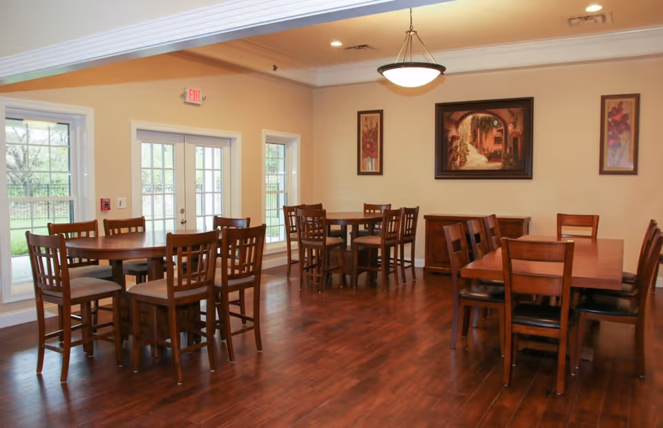 A dining room with several wooden tables and chairs arranged neatly on a polished wooden floor. The room has beige walls adorned with three framed paintings, and large windows and glass doors letting in natural light. A ceiling light fixture hangs in the center of the room.