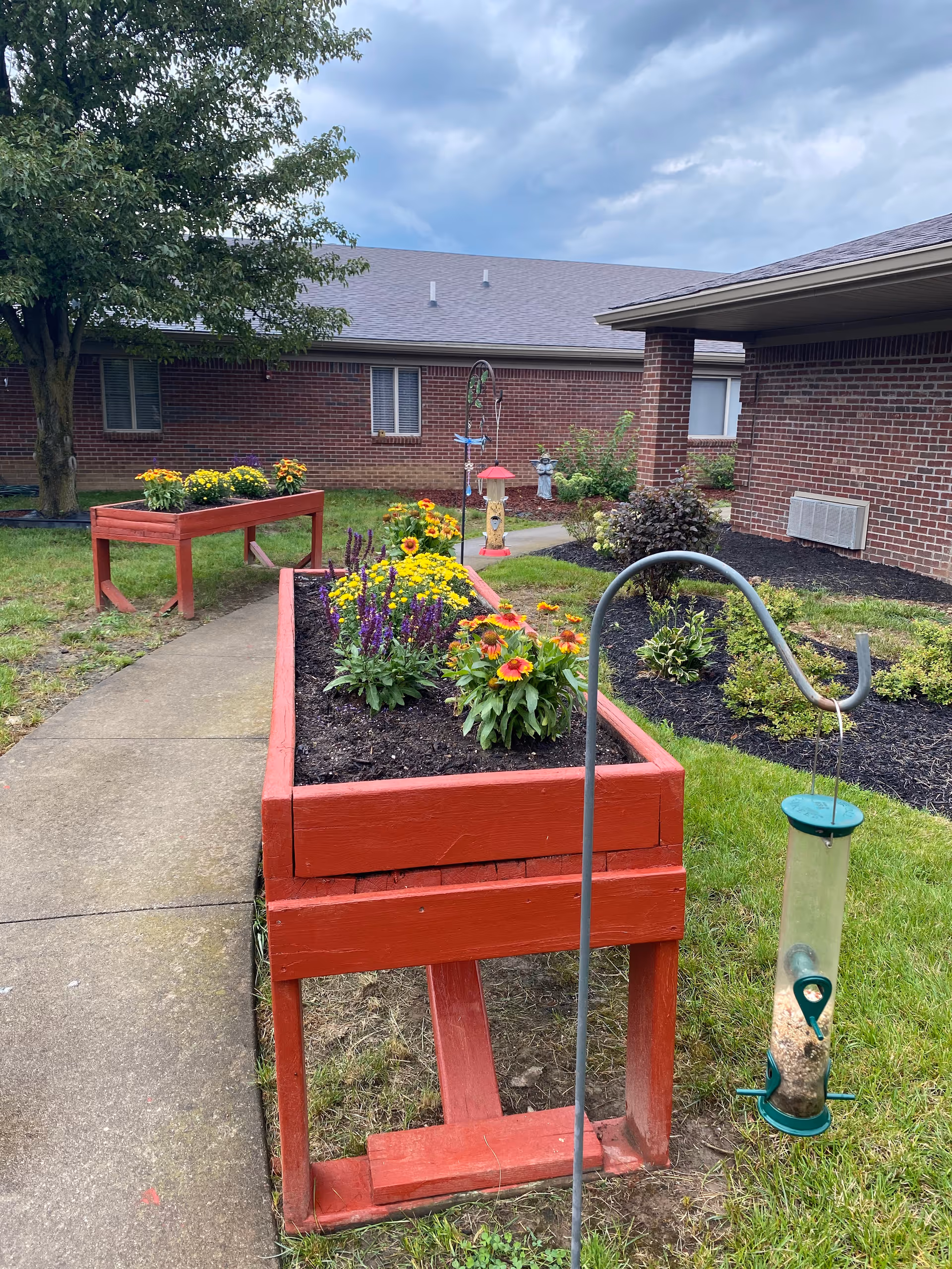 Outdoor garden area at Majestic Care of North Vernon featuring raised red wooden flower beds filled with colorful flowers, a bird feeder hanging from a metal hook, a concrete pathway, and a brick building in the background under a cloudy sky.