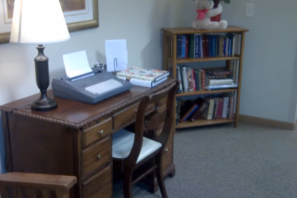 A small office or study area with a wooden desk and chair. On the desk, there is a lamp, a typewriter with paper, and a few books. In the background, there is a wooden bookshelf filled with books and a small stuffed animal on top.