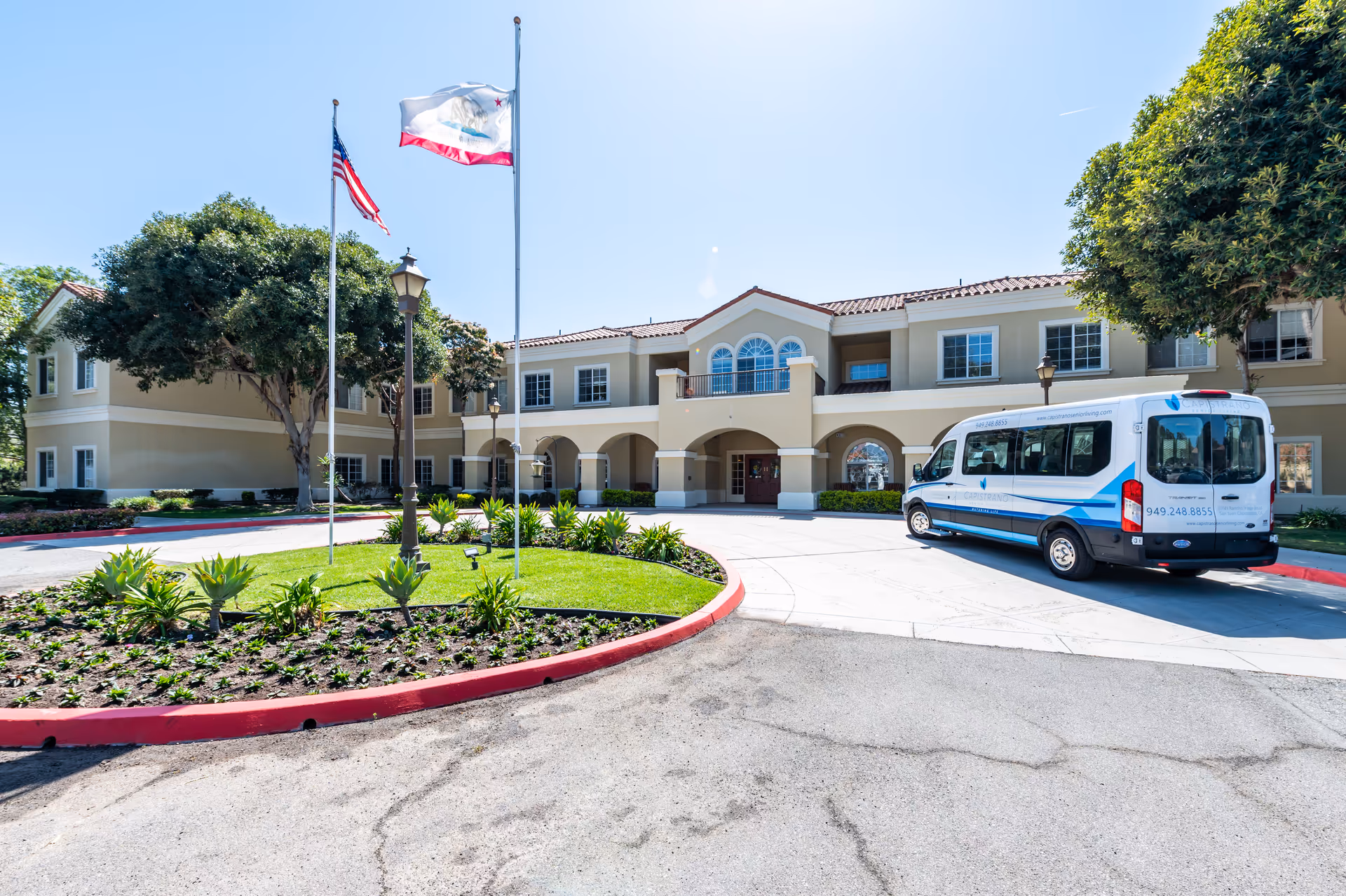 Front exterior view of Capistrano Senior Living facility with a circular driveway, landscaped garden area with plants and two flagpoles flying the American and California state flags. A white and blue shuttle van is parked near the entrance under a clear blue sky.