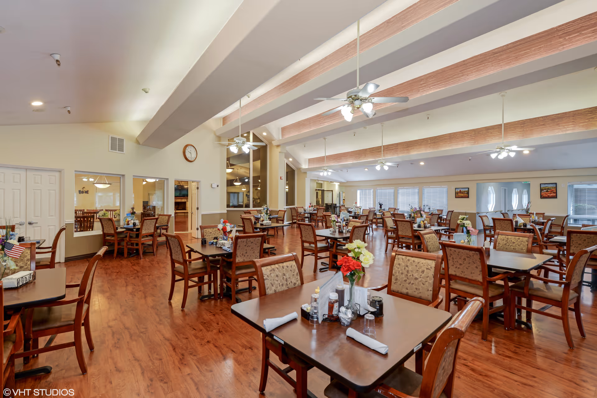 Spacious dining hall with wooden tables, chairs, and centerpieces in a senior living facility.