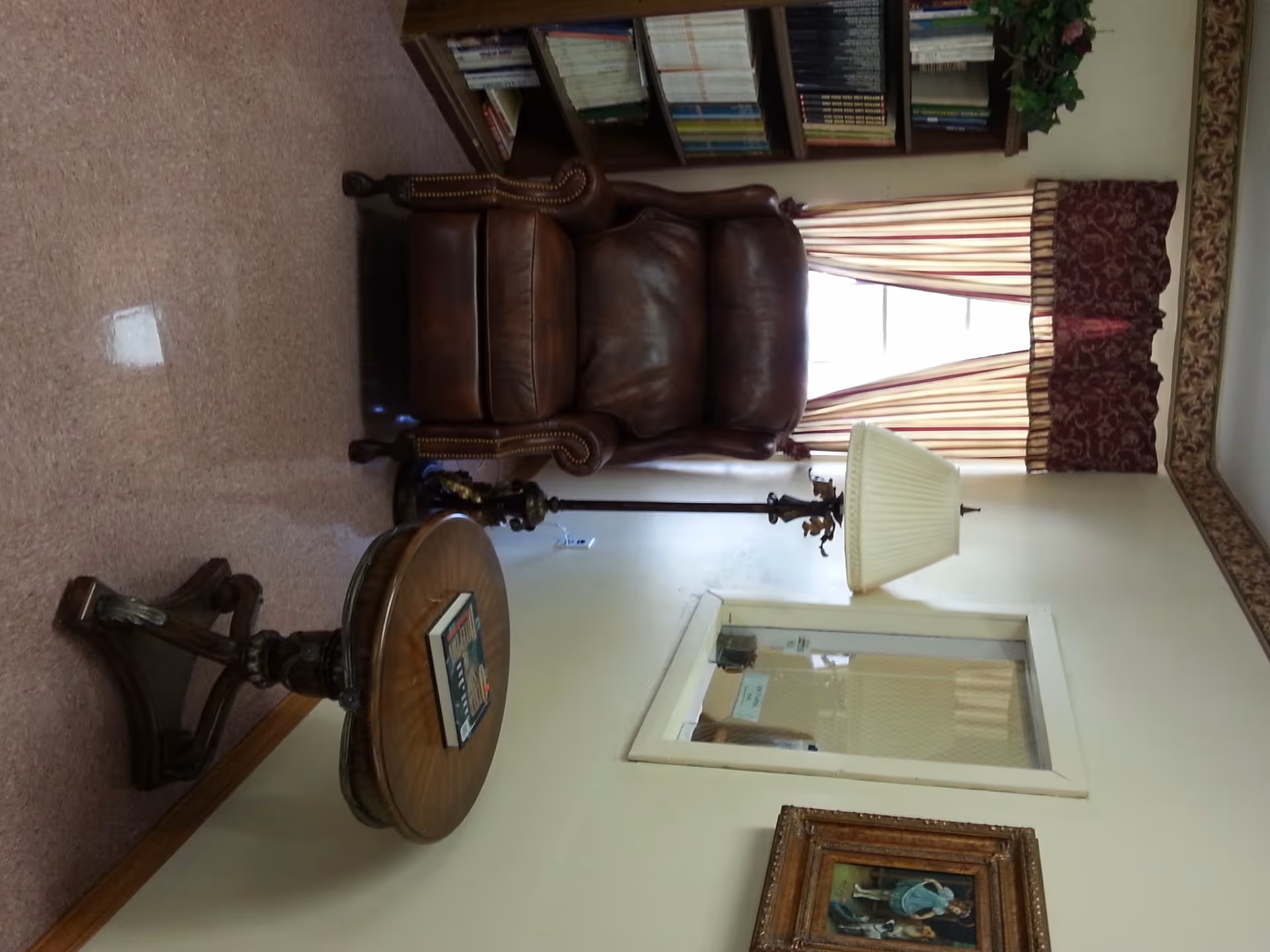 A cozy corner of a room featuring a brown leather armchair, a round wooden side table with a book on it, a tall floor lamp with a cream-colored shade, a window with striped curtains and a decorative valance, a bookshelf filled with books, and a framed painting on the wall.