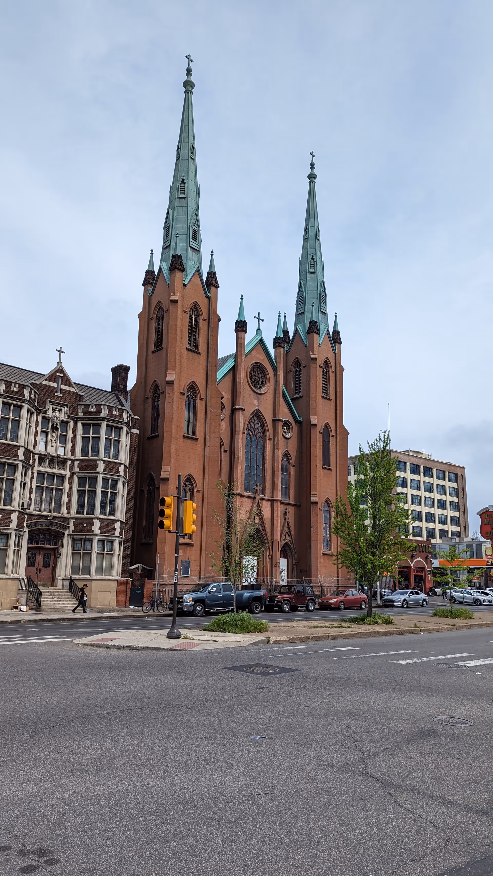 Brick church with tall green spires on a city street, cars nearby.