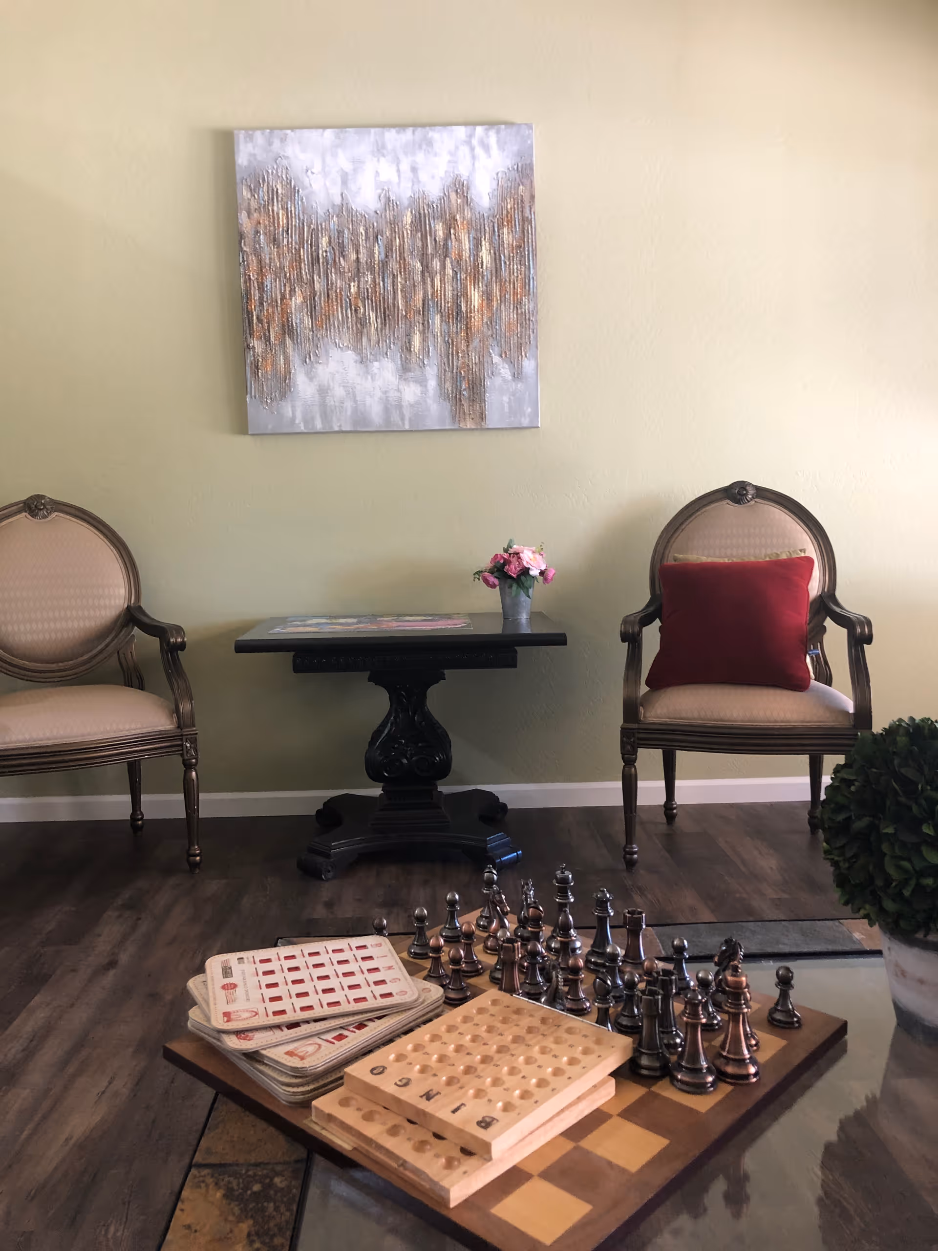 A cozy seating area with two upholstered chairs and a small table under abstract wall art, and a coffee table in front holding a chess set and game boards.