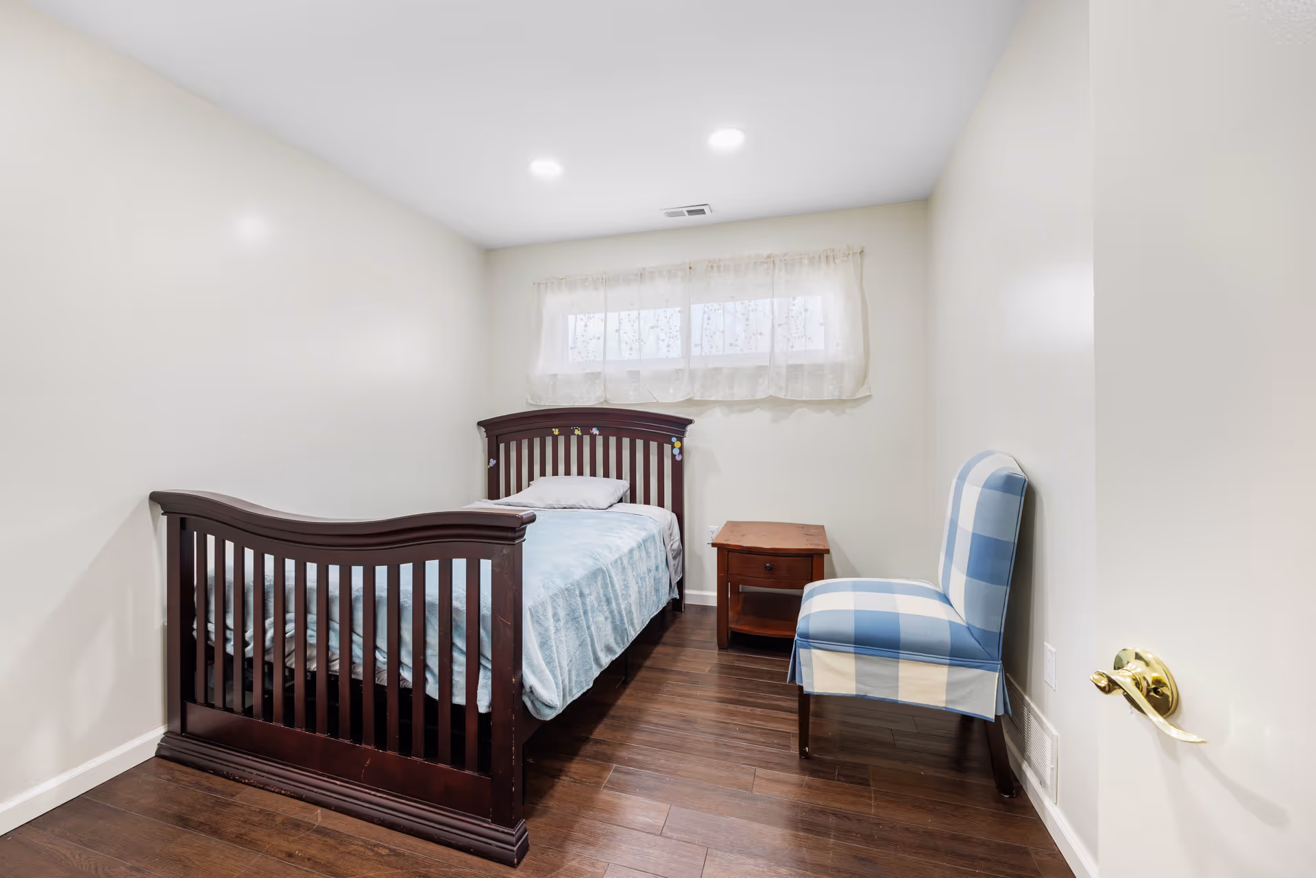 A small bedroom with a dark wooden bed, nightstand, window, and blue plaid chair.