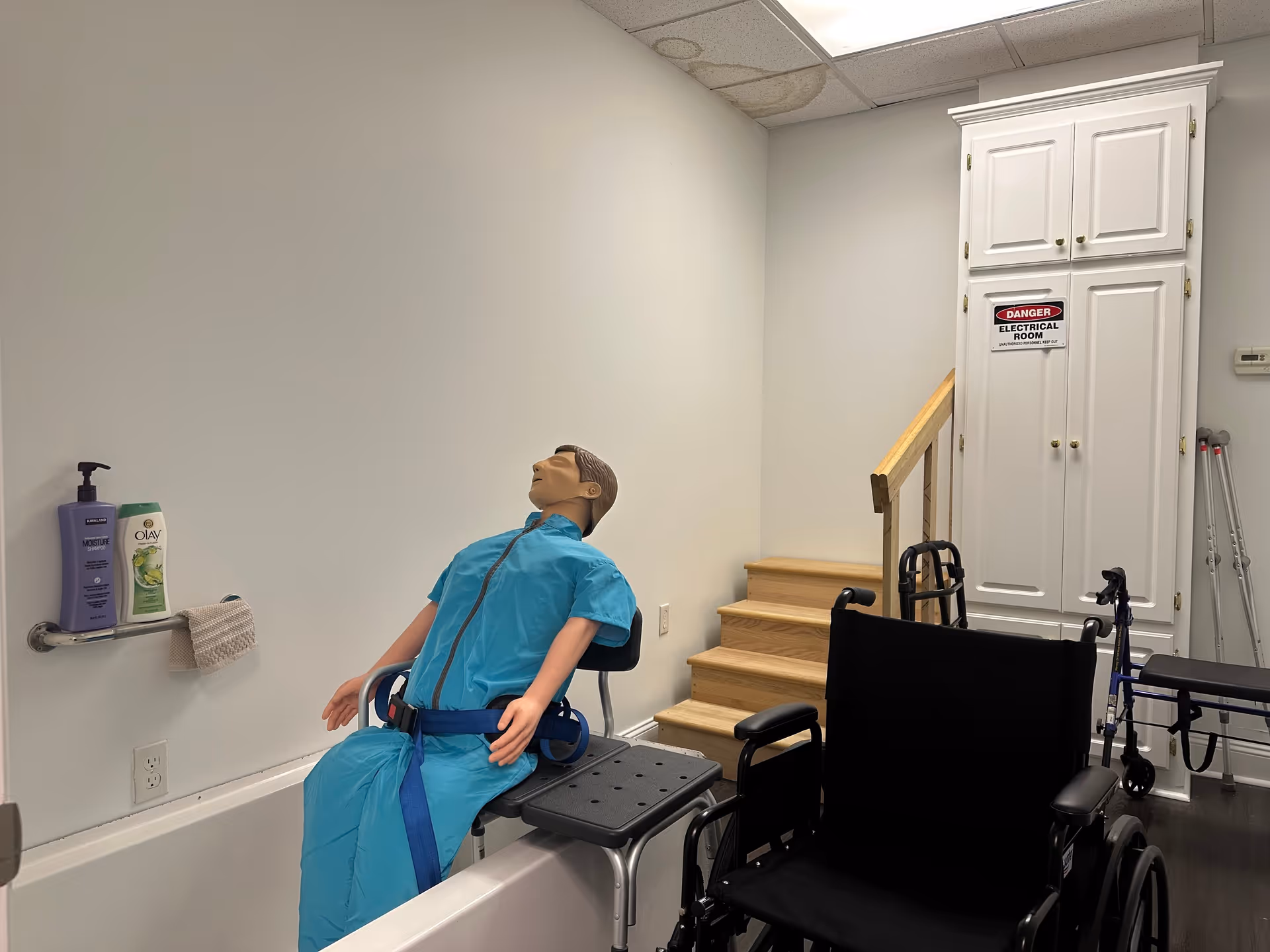 Medical training mannequin in blue scrubs sits beside a wheelchair in a clinical room.
