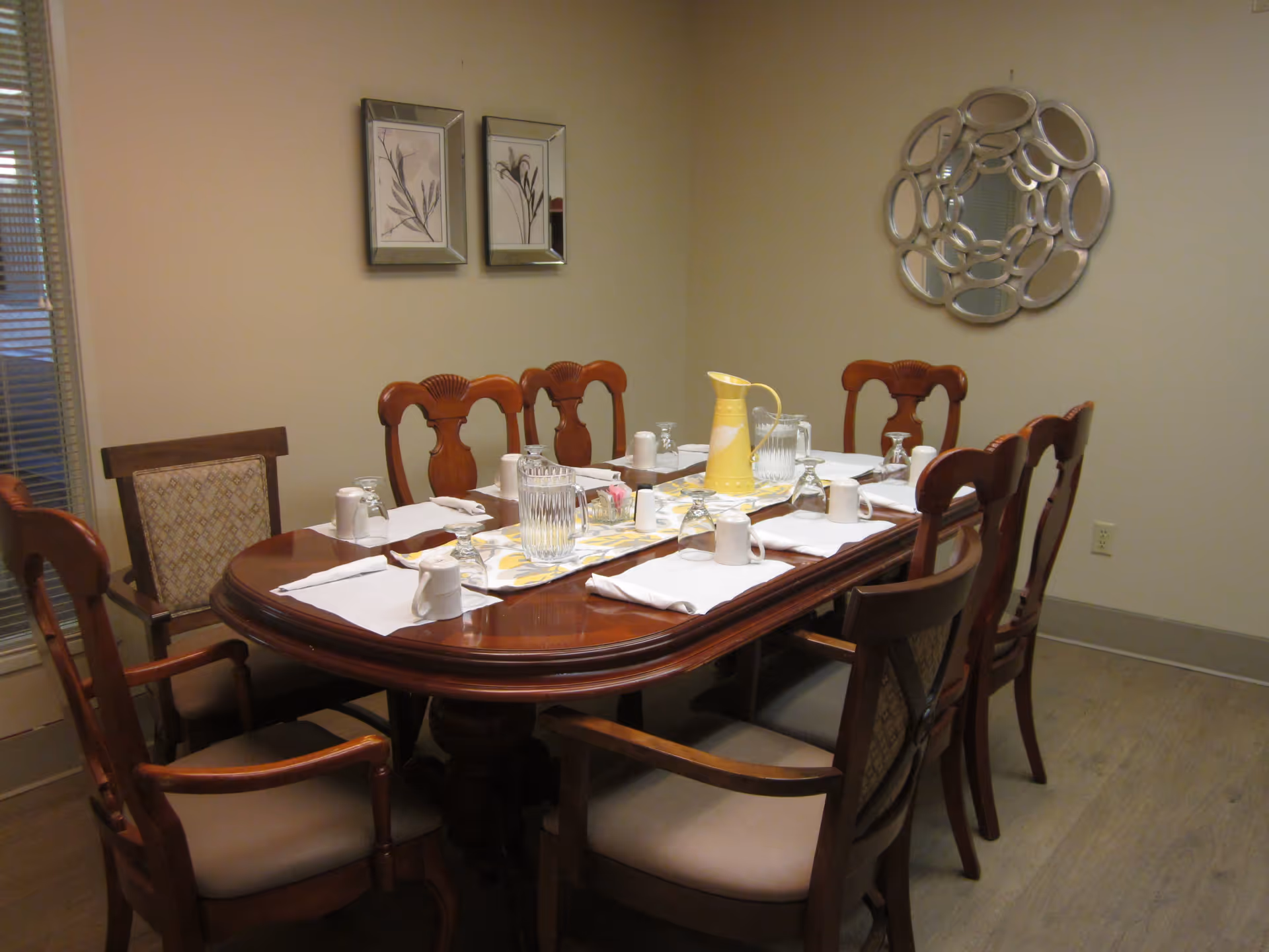 A dining room with a polished wooden table set for eight people. The table has white napkins, upside-down glasses, white mugs, a yellow pitcher, and a clear water pitcher on a decorative table runner. The room has beige walls with two framed botanical prints and a decorative circular mirror. There are wooden chairs with cushioned seats around the table.