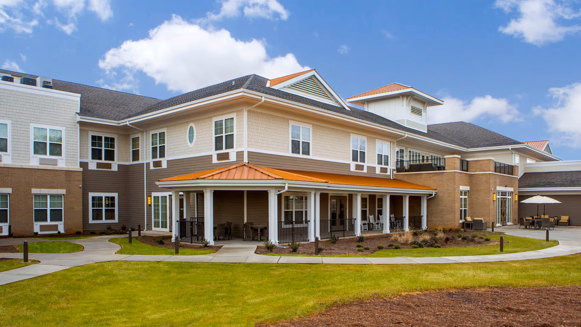 Two-story senior living building with a covered porch, paved walkways and a landscaped lawn under a blue sky.