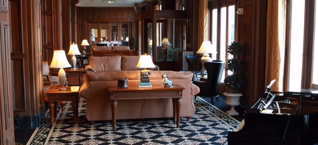A spacious, elegant living room with wooden paneling, large windows with curtains, a patterned carpet, multiple table lamps, a grand piano, and comfortable seating including a sofa and armchairs.