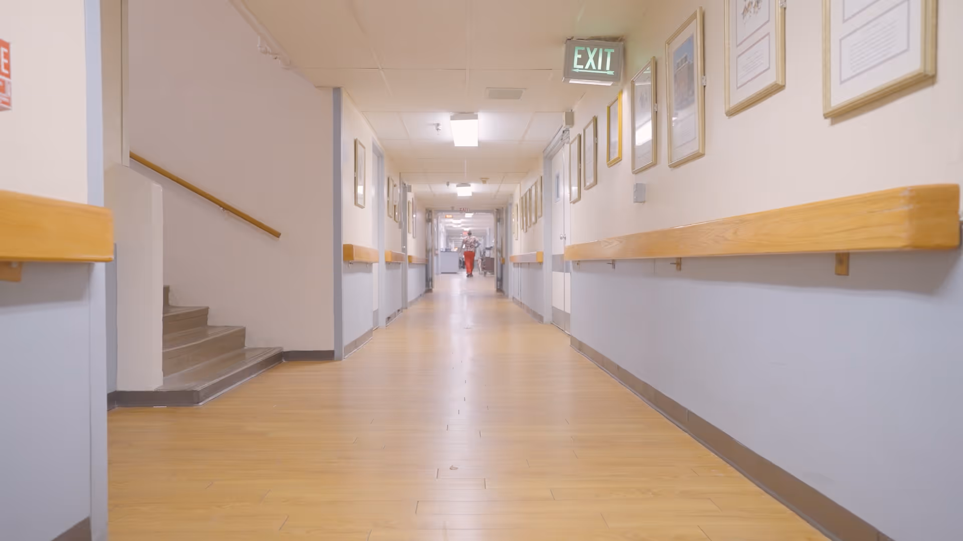 Long well-lit corridor in a senior care facility with wooden handrails, framed pictures, and an exit sign.
