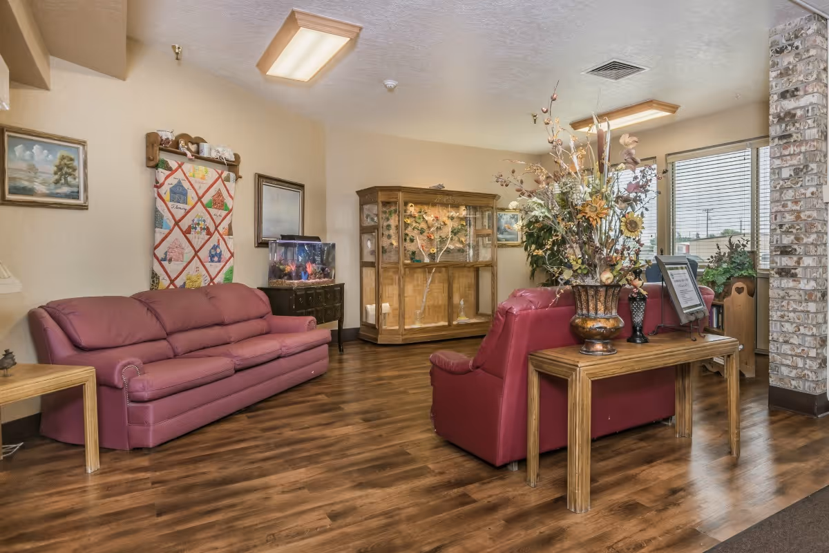 A cozy living room area in a nursing home with two maroon leather sofas, a wooden side table with a large floral arrangement, a wooden display cabinet with decorative items, an aquarium on a small table, framed paintings on the walls, and large windows with blinds allowing natural light in.