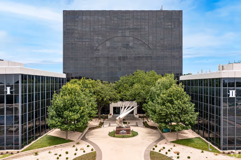 A modern glass-front building with a central sculpture and trees in a courtyard.