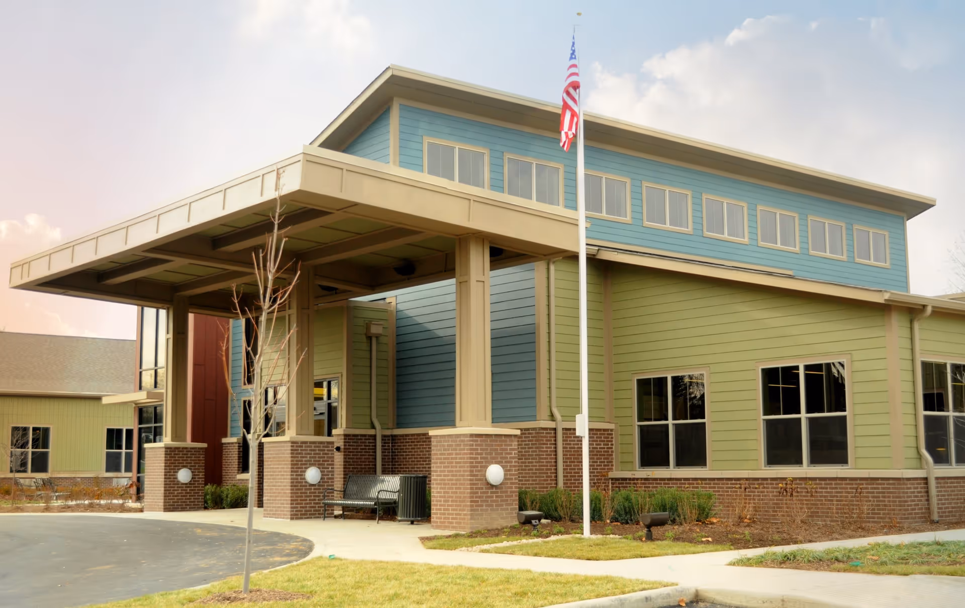 Front entrance of a modern senior living building with a covered porte-cochere, American flagpole, and green and blue siding.