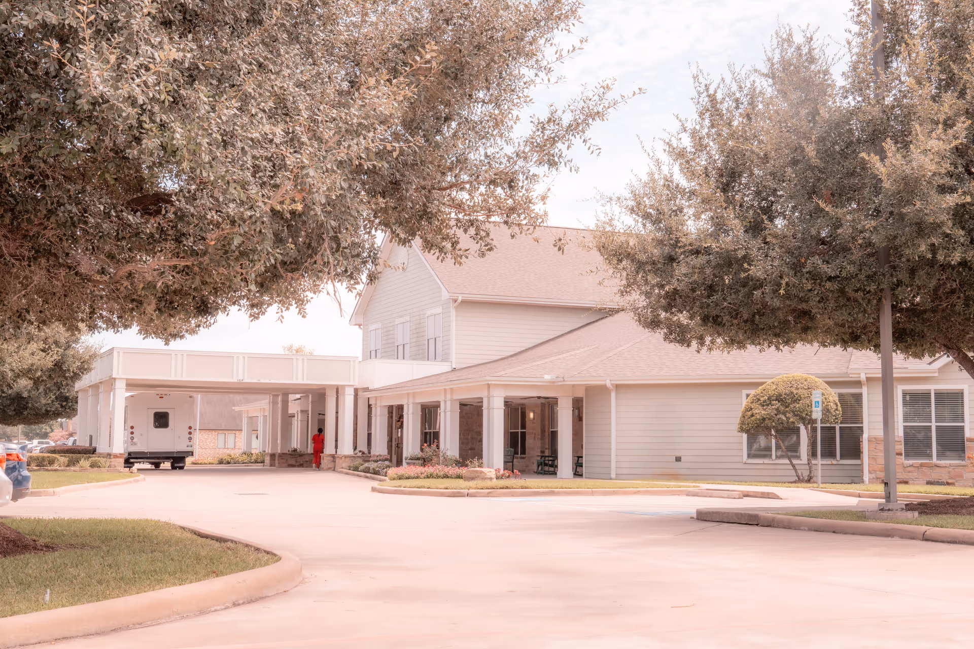 Front exterior of a two-story assisted living facility with a covered entrance, driveway, and surrounding trees.