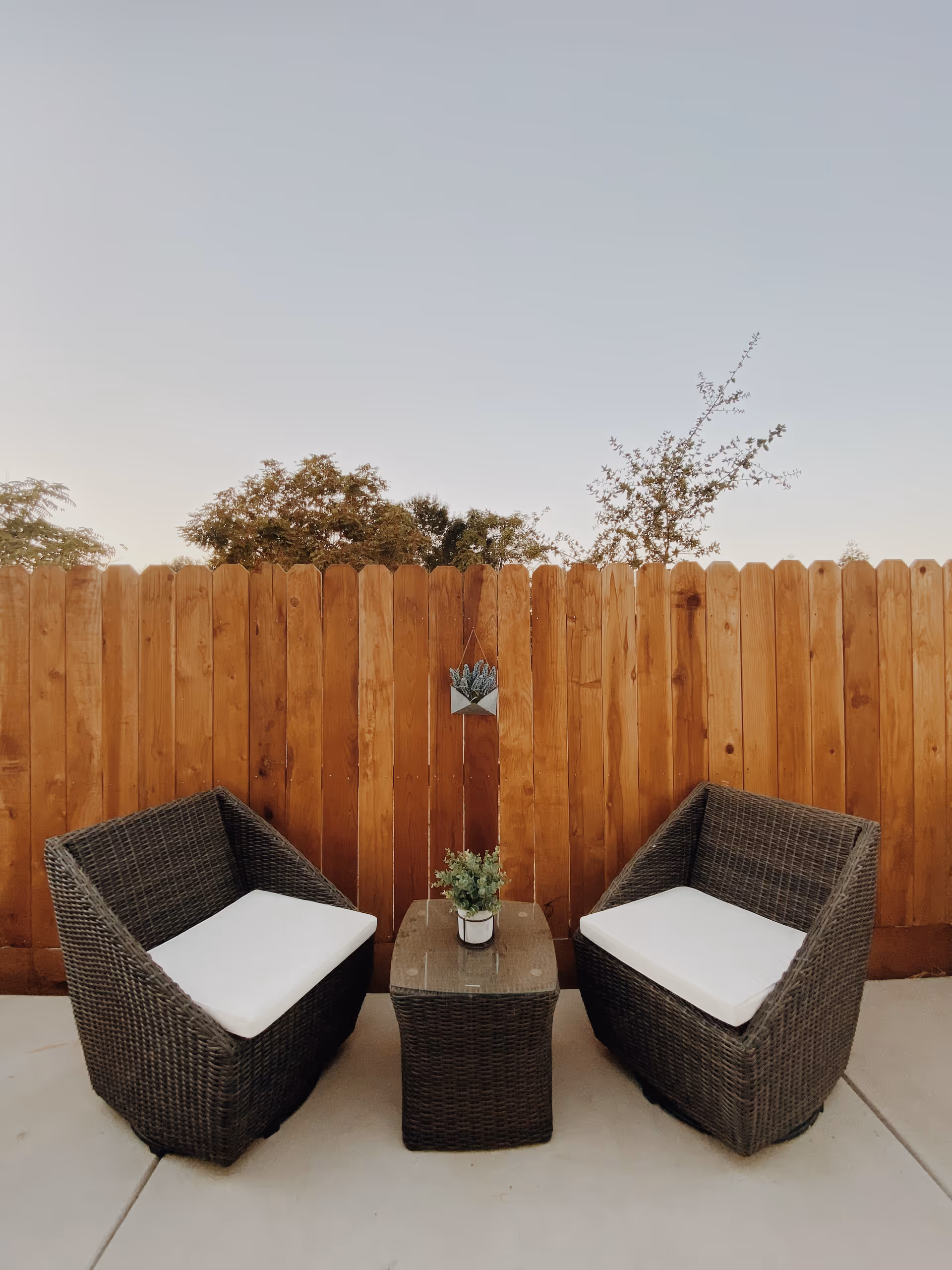 Two dark wicker chairs with white cushions and a matching wicker table with a small potted plant on top, set on a concrete patio in front of a wooden fence with some trees visible above the fence under a clear sky.