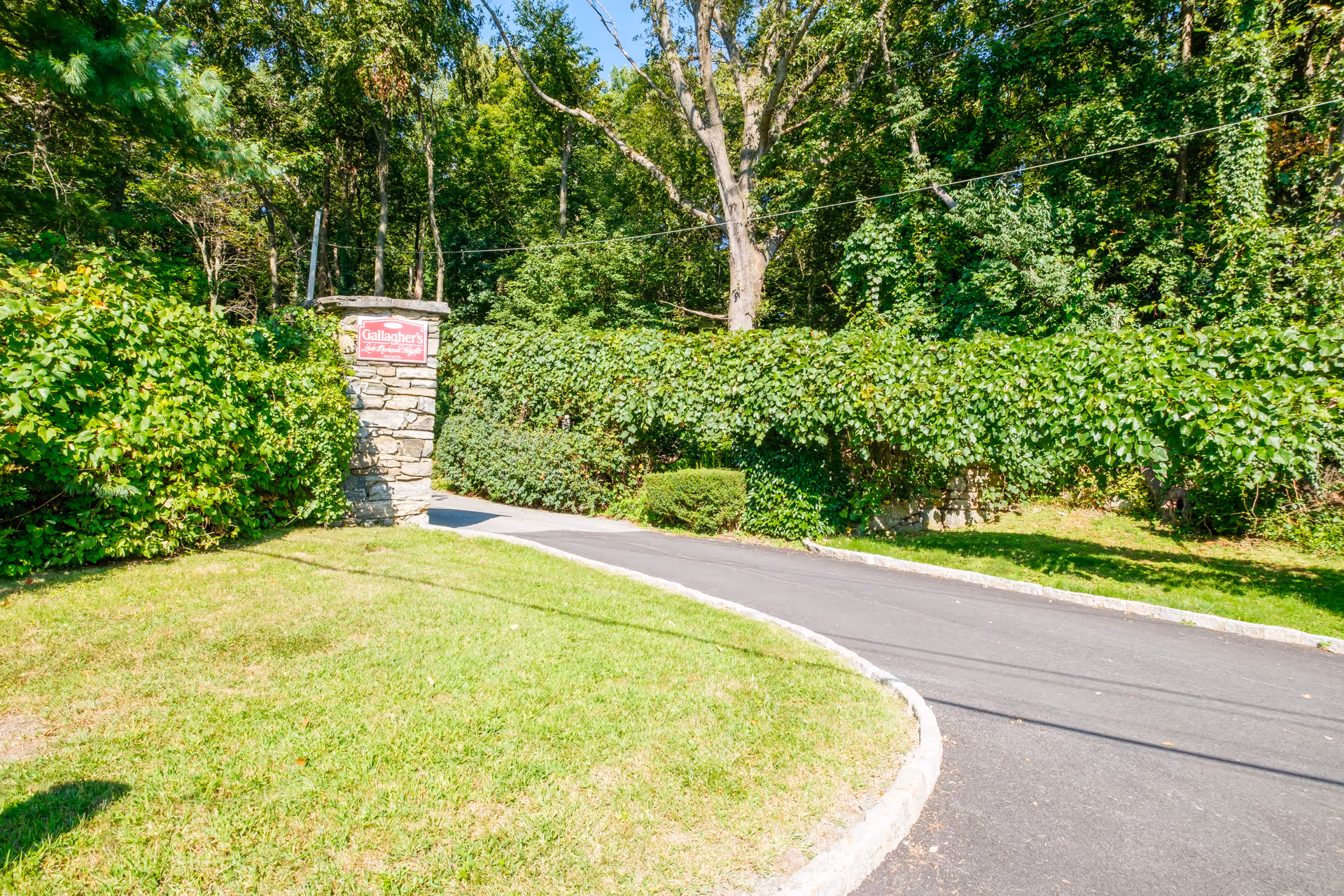 Curving paved driveway leading past a stone entrance pillar with a 'Gallagher' sign surrounded by dense trees and shrubs.