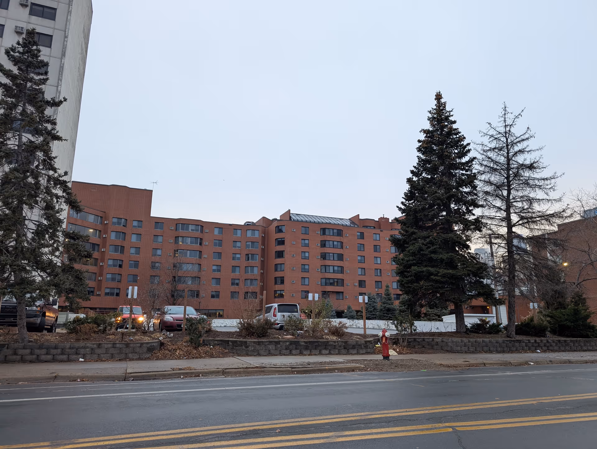 Wide view of a multi-story red-brick apartment building with trees, parked cars, and a street in the foreground.