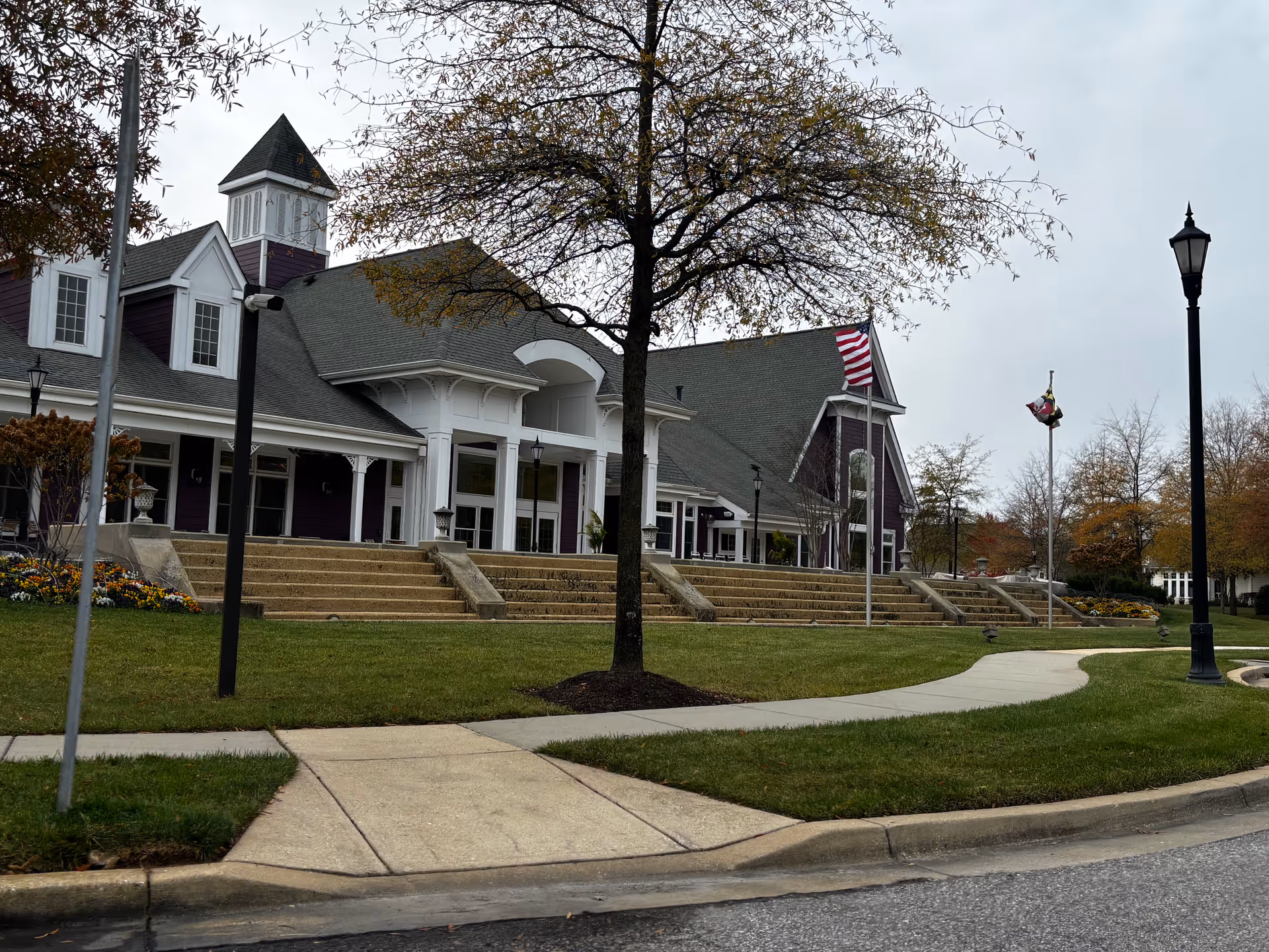 Exterior view of a senior living facility building with a large porch, steps leading up to the entrance, and two flagpoles displaying the American flag and another flag. The building has a gray roof, white trim, and purple siding. There is a tree and a curved sidewalk in the foreground with street lamps and landscaped grass and flowers.