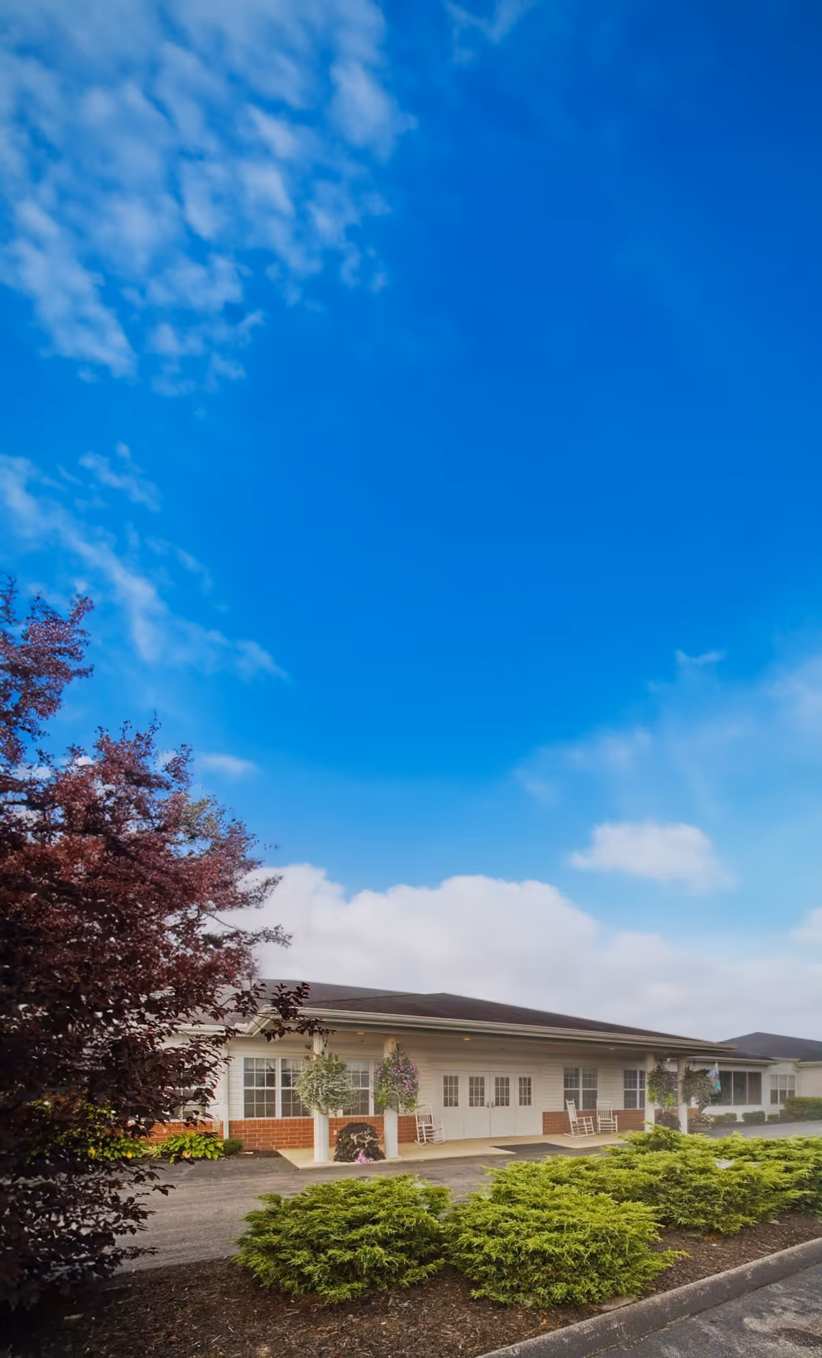 Exterior view of a single-story building with white siding and a dark roof under a bright blue sky with some clouds. The building has multiple windows and doors, with potted plants and rocking chairs on the porch area. There are green shrubs and a tree with reddish leaves in the foreground.