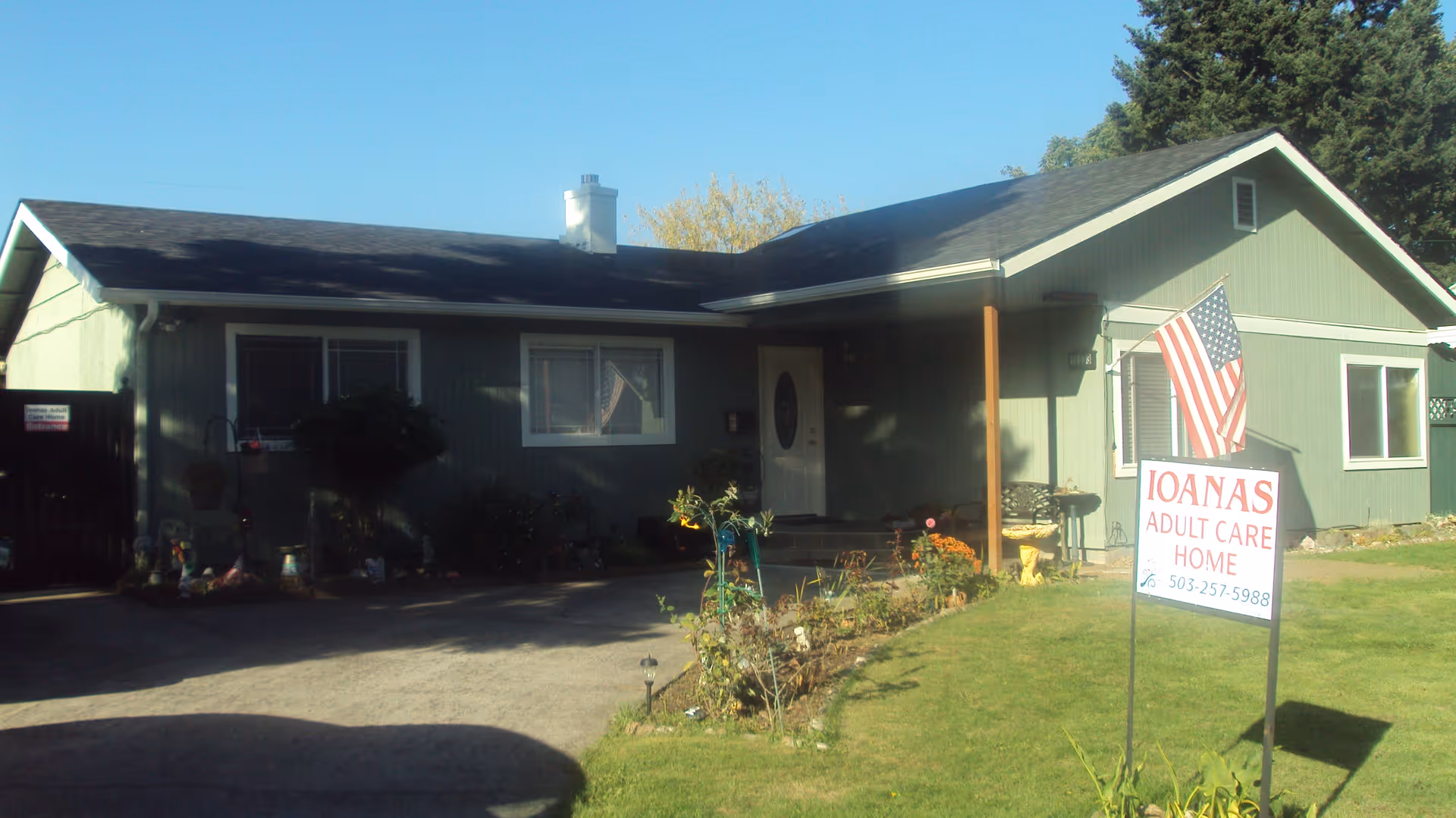 Single-story green house with a front porch, an American flag, and a sign in the yard that reads 'Ioanas Adult Care Home' with a phone number. The house has a driveway and some plants and flowers in front.