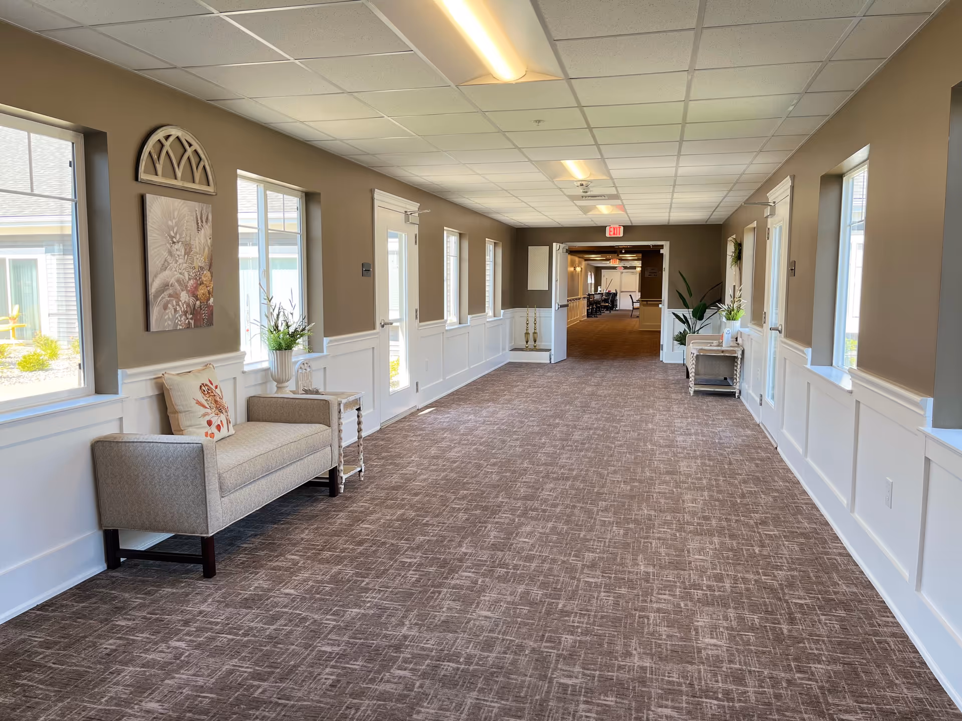 Wide hallway in a senior living facility with seating, plants, and windows.