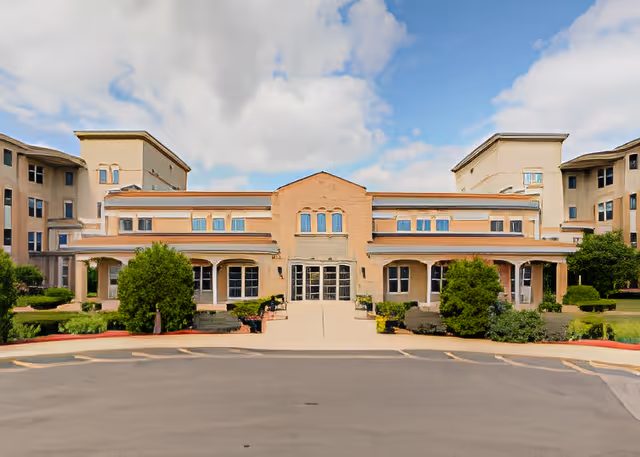 Front entrance and facade of a multi-story beige senior living facility with landscaped shrubs and a driveway.