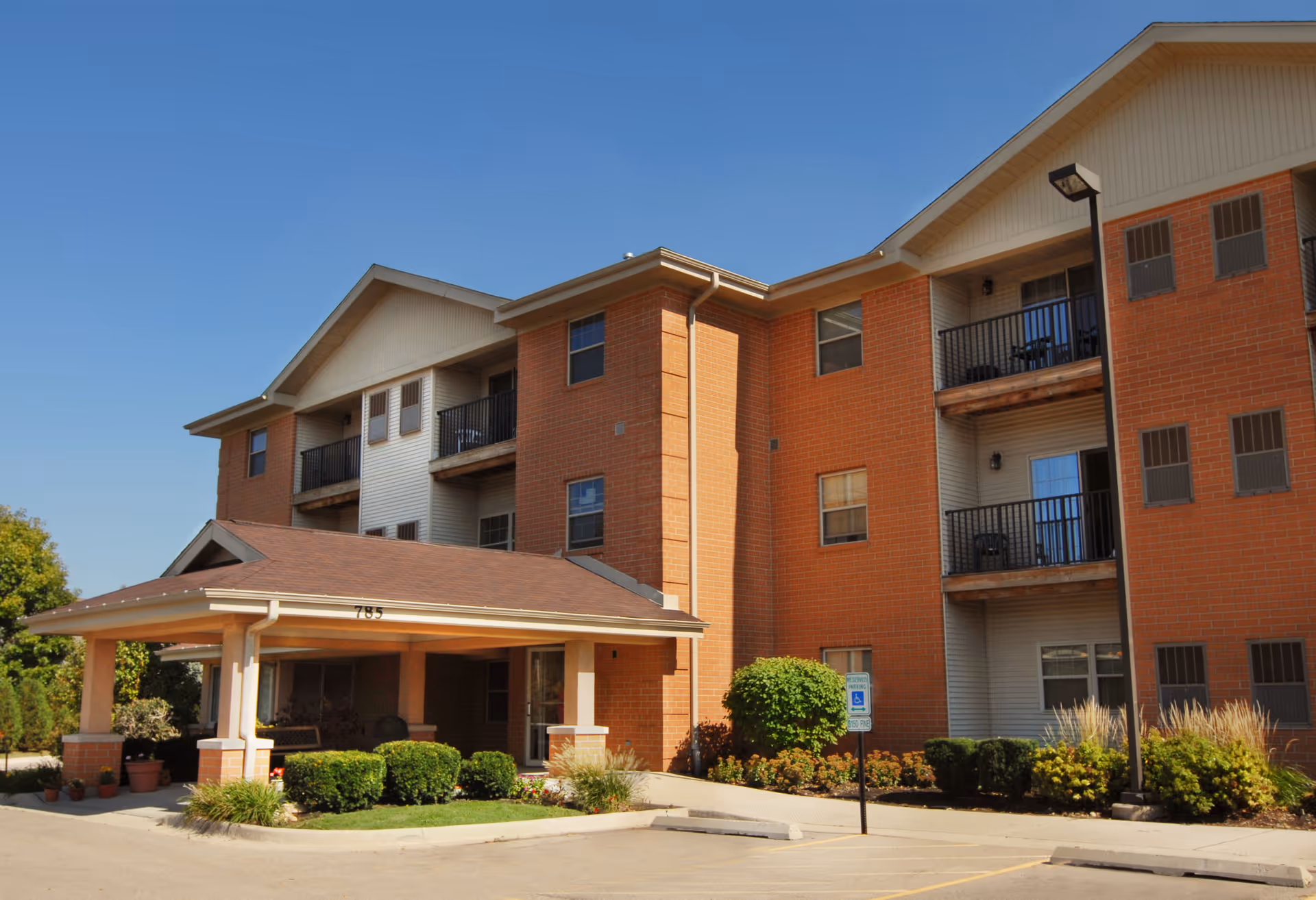 Three-story brick residential building with a covered entrance, balconies and landscaped front under a clear blue sky.