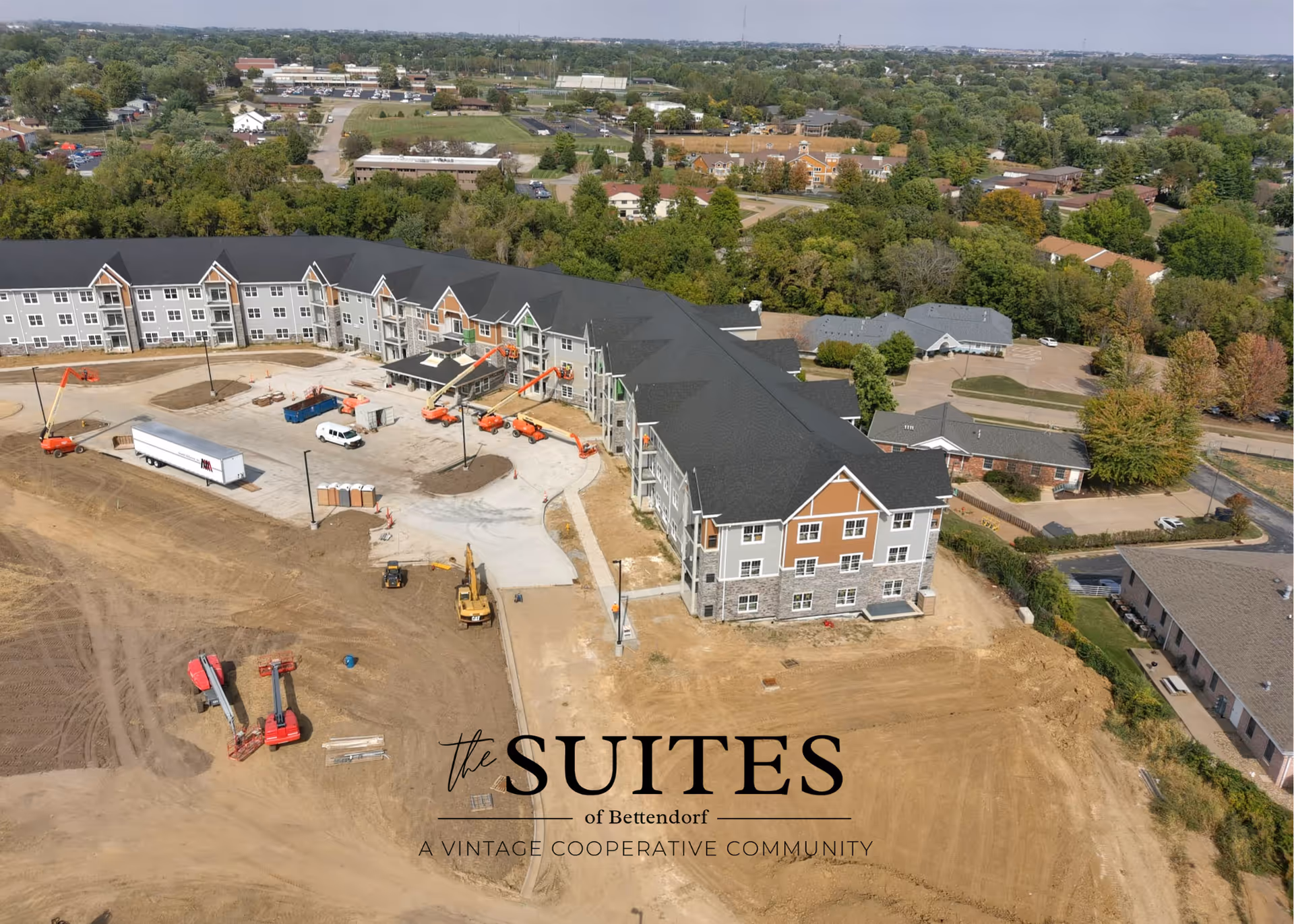 Aerial view of a large, multi-story building under construction surrounded by dirt and construction equipment, with trees and other buildings in the background.