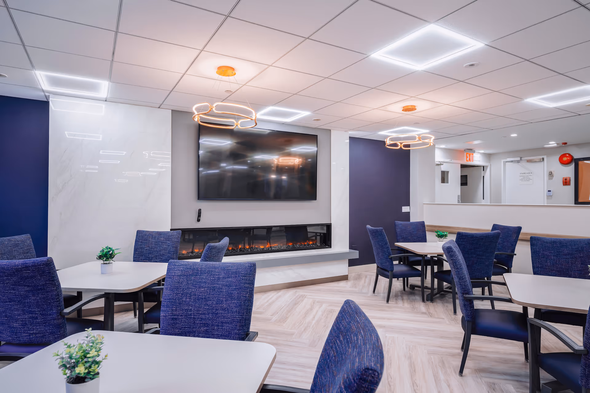 A modern common area with several tables and blue upholstered chairs arranged around them. The room features a large flat-screen TV mounted above a sleek electric fireplace set into a wall with white and dark blue panels. Two contemporary circular light fixtures hang from the ceiling, and small potted plants are placed on the tables. The floor has a light wood pattern, and there is an exit door visible in the background.
