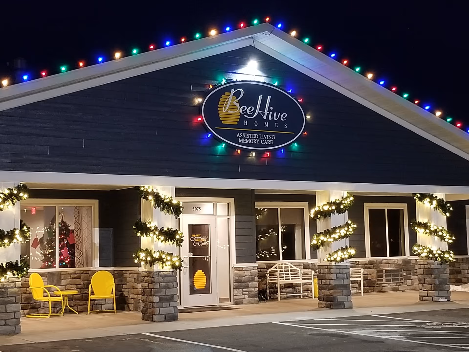 Night exterior of BeeHive Homes facility with holiday lights, chairs outside entrance.