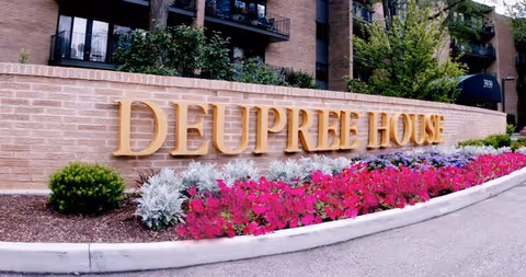 Exterior view of Deupree House entrance with brick wall, gold letters, and vibrant flowerbeds.