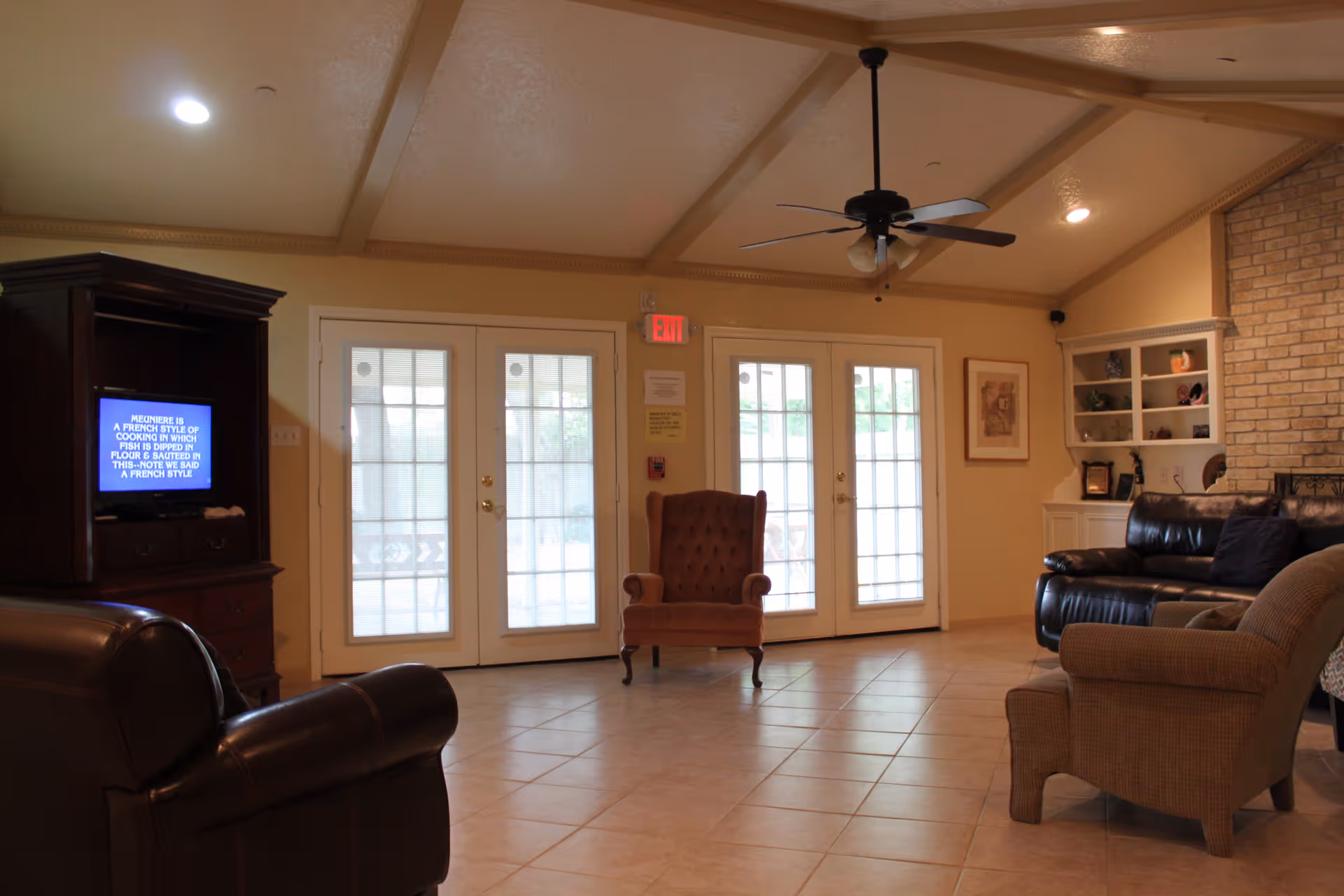 A cozy living room with tiled floor, featuring a brown leather armchair, a beige upholstered armchair, a brown leather sofa, and a single brown upholstered armchair in the center. There is a wooden cabinet with a TV on the left side, and built-in white shelves with decorative items on the right. The room has a ceiling fan with lights and two sets of glass double doors leading outside.