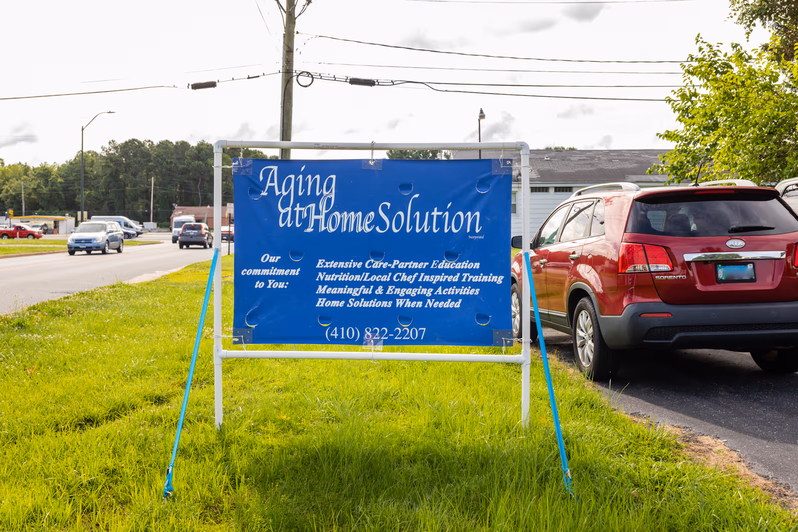 Blue Aging at Home Solution sign on a metal frame in a grassy roadside lot.
