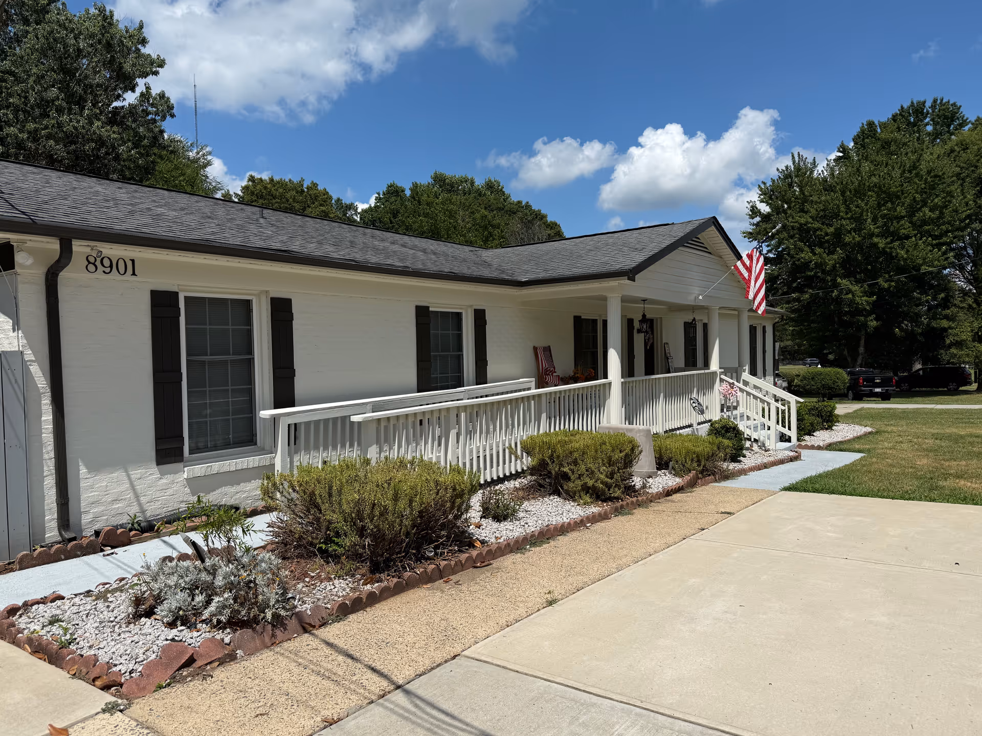 Front view of a single-story white building with a porch, railing, landscaping, and an American flag.