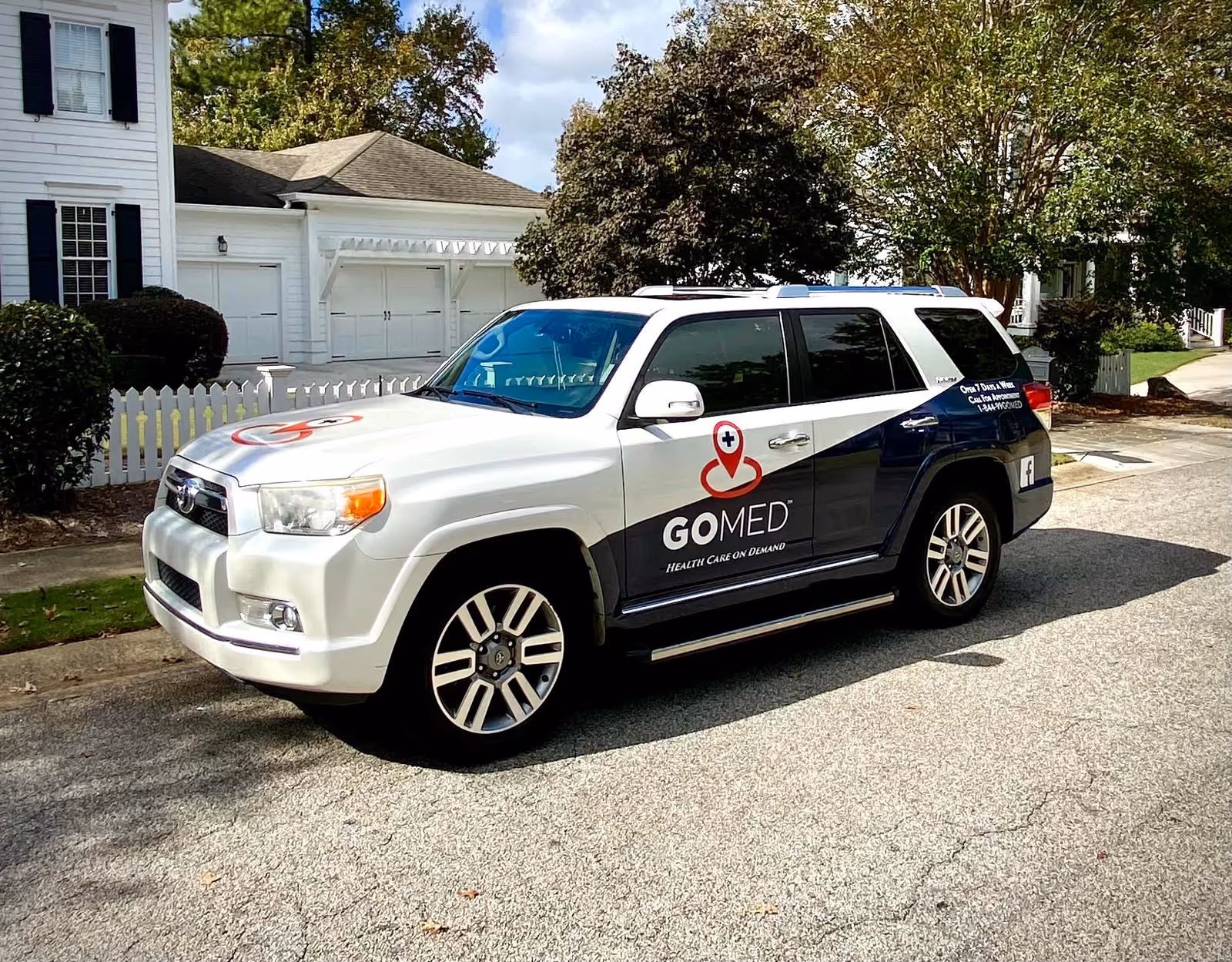 White GMC SUV with GOMED logo parked on a suburban street in front of white houses.