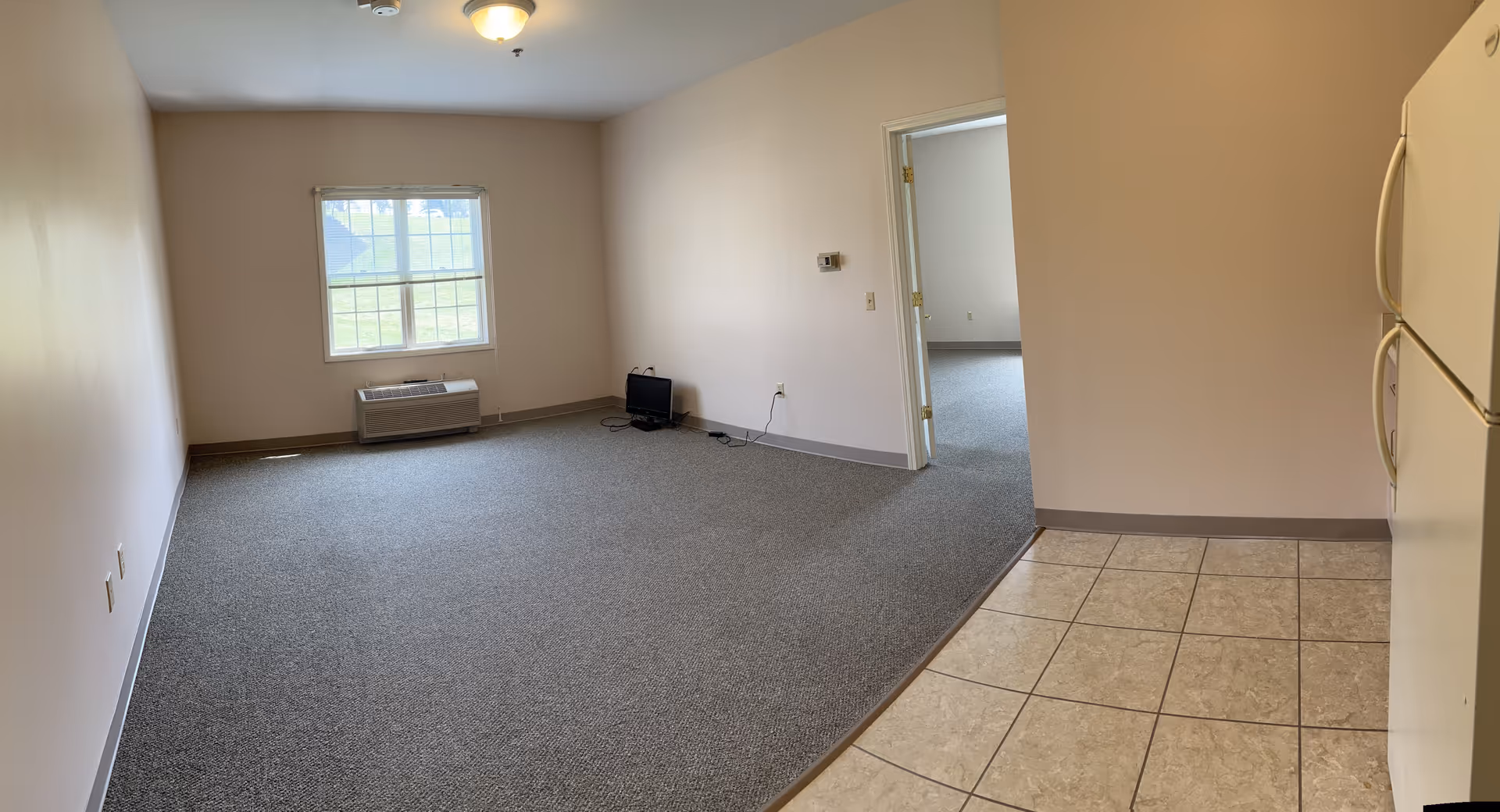 Spacious, empty interior living area with window, air conditioner, and doorway to another room.