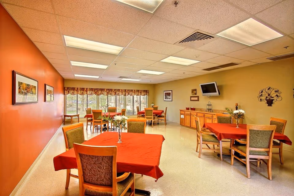 A well-lit dining room with red tablecloths and wooden chairs in a senior facility.