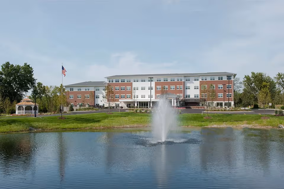 A large multi-story senior living facility building named Oak Hill with a brick and white exterior, situated behind a pond with a water fountain in the center. There is a gazebo to the left and an American flag on a flagpole near the building. The scene is set on a clear day with trees and green grass surrounding the area.