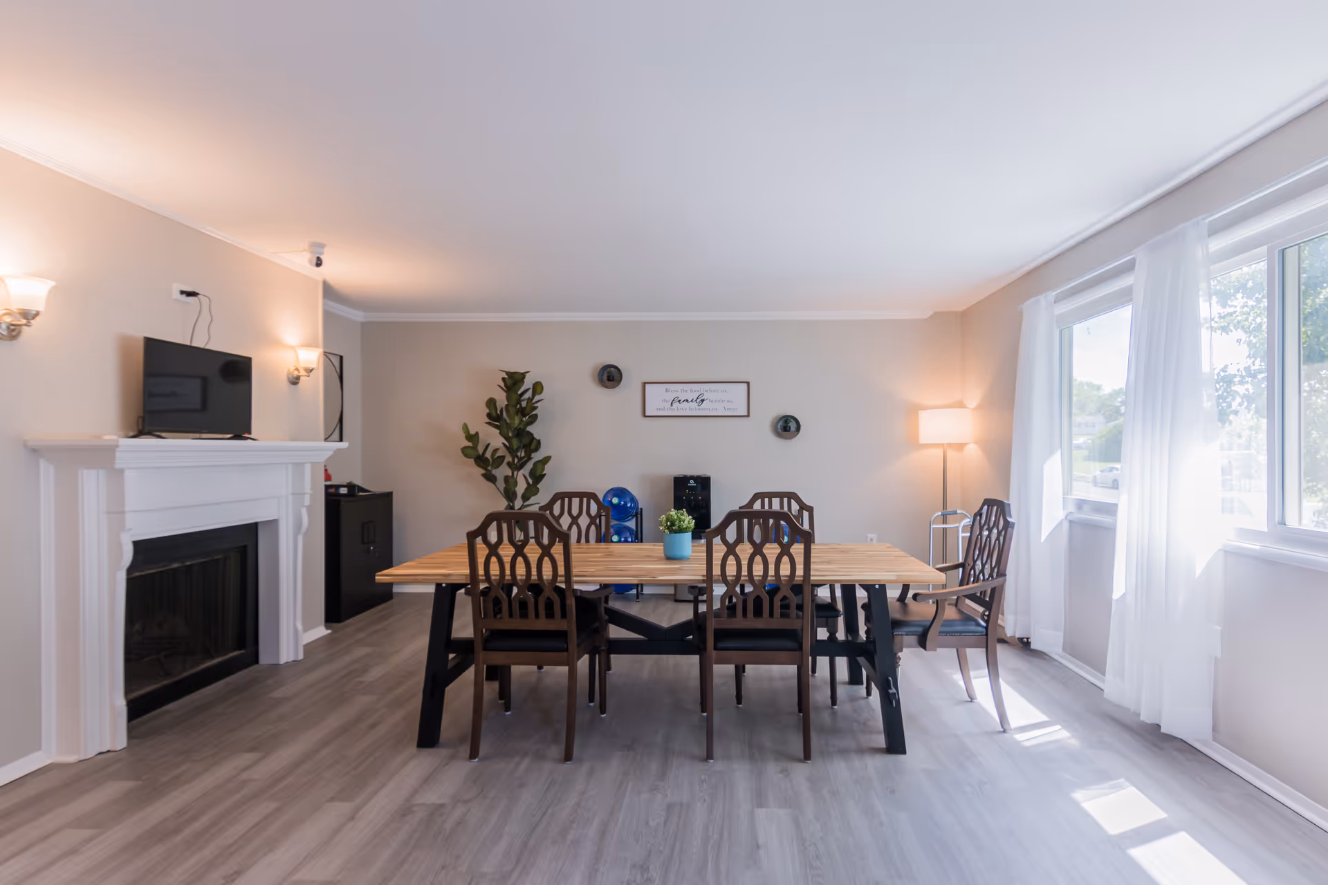 Bright dining room with a wooden table, eight chairs, windows with white curtains, and a fireplace.