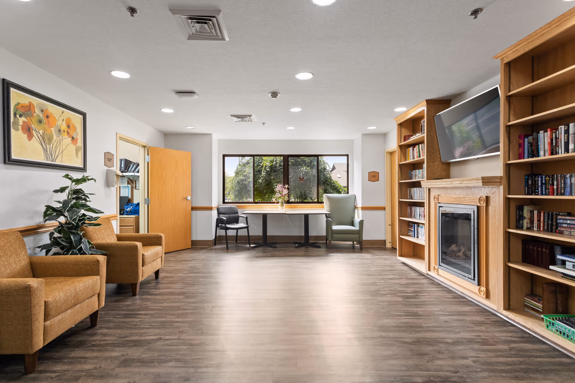 A bright and spacious common area with wooden flooring, two brown armchairs on the left, a green armchair and a black chair near a window with a table in between. On the right side, there are built-in wooden bookshelves surrounding a fireplace with a mounted flat-screen TV above it. A framed floral painting and a potted plant are visible on the left wall.
