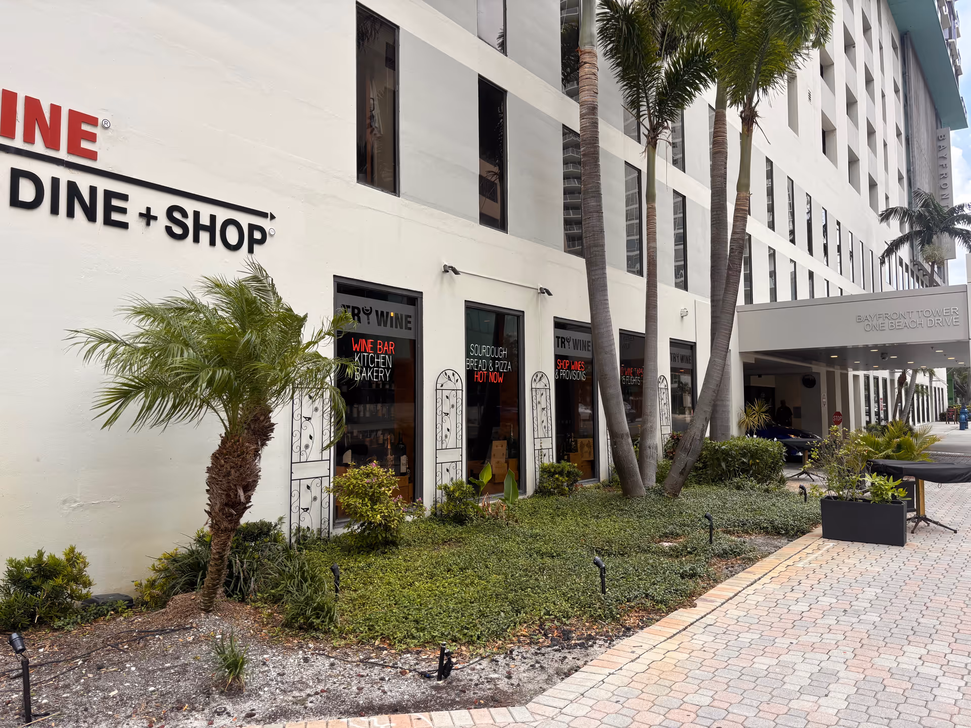 White building exterior with DINE and SHOP signs, palm trees, and a paved walkway.
