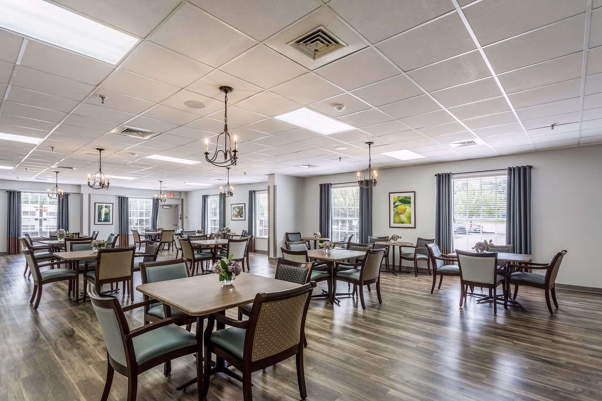 A spacious dining room with multiple wooden tables and chairs arranged neatly. Each table has a small floral centerpiece. The room has large windows with gray curtains, framed artwork on the walls, and chandeliers hanging from a white tiled ceiling. The floor is covered with wood-style laminate.
