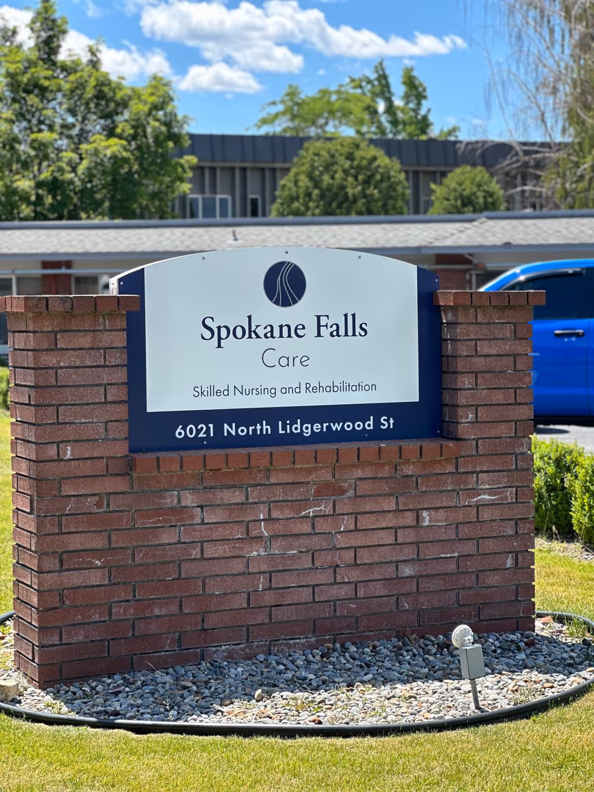 Brick sign reading Spokane Falls Care outside a facility with trees and gravel landscaping.