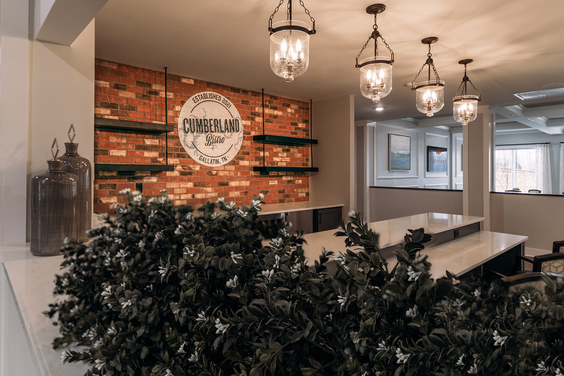 Interior view of a dining area with a brick accent wall featuring a round sign that reads 'Cumberland Bistro, Established 2021, Gallatin, TN'. The room has a long white countertop with green plants in the foreground, hanging glass pendant lights, and framed artwork on the walls in the background.